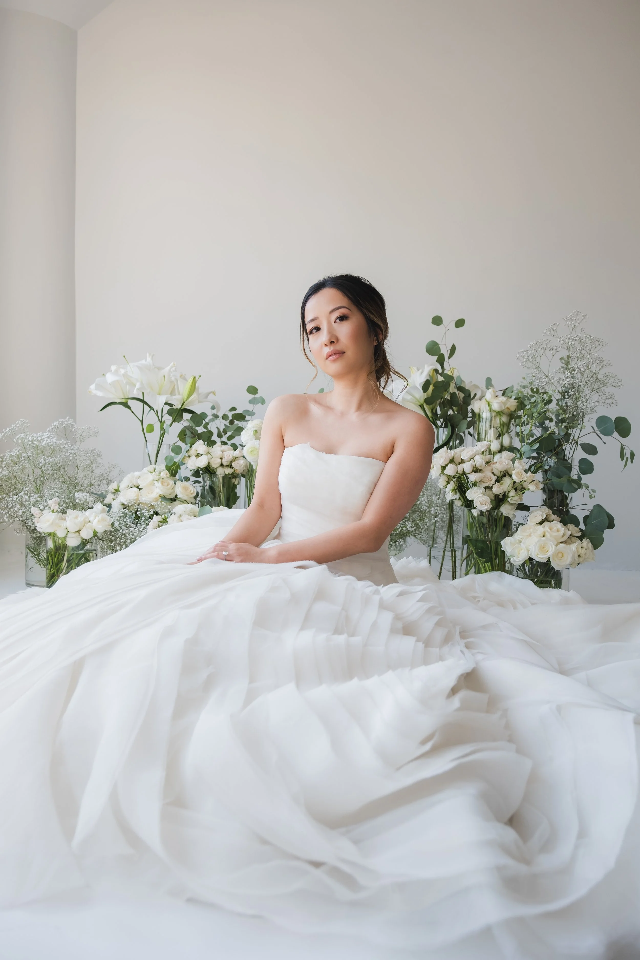 A woman in a white wedding dress sitting in front of white flowers arranged in vases.