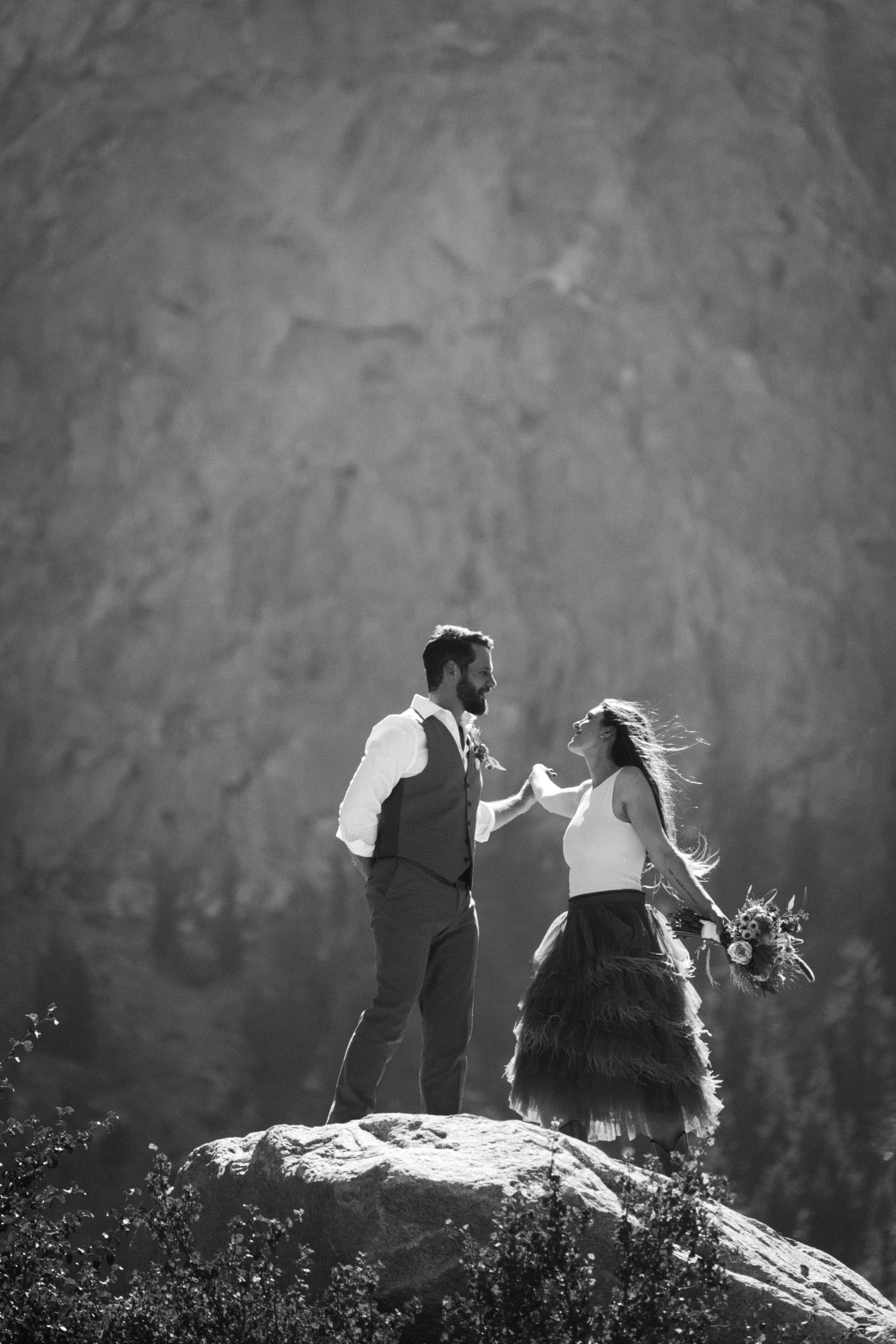 A couple stands on a rock outdoors, with trees in the background, sharing a moment on their wedding day. The groom has a beard and is in a vest and white shirt, holding the bride's hand. The bride has long hair, wears a white top and a layered skirt,
