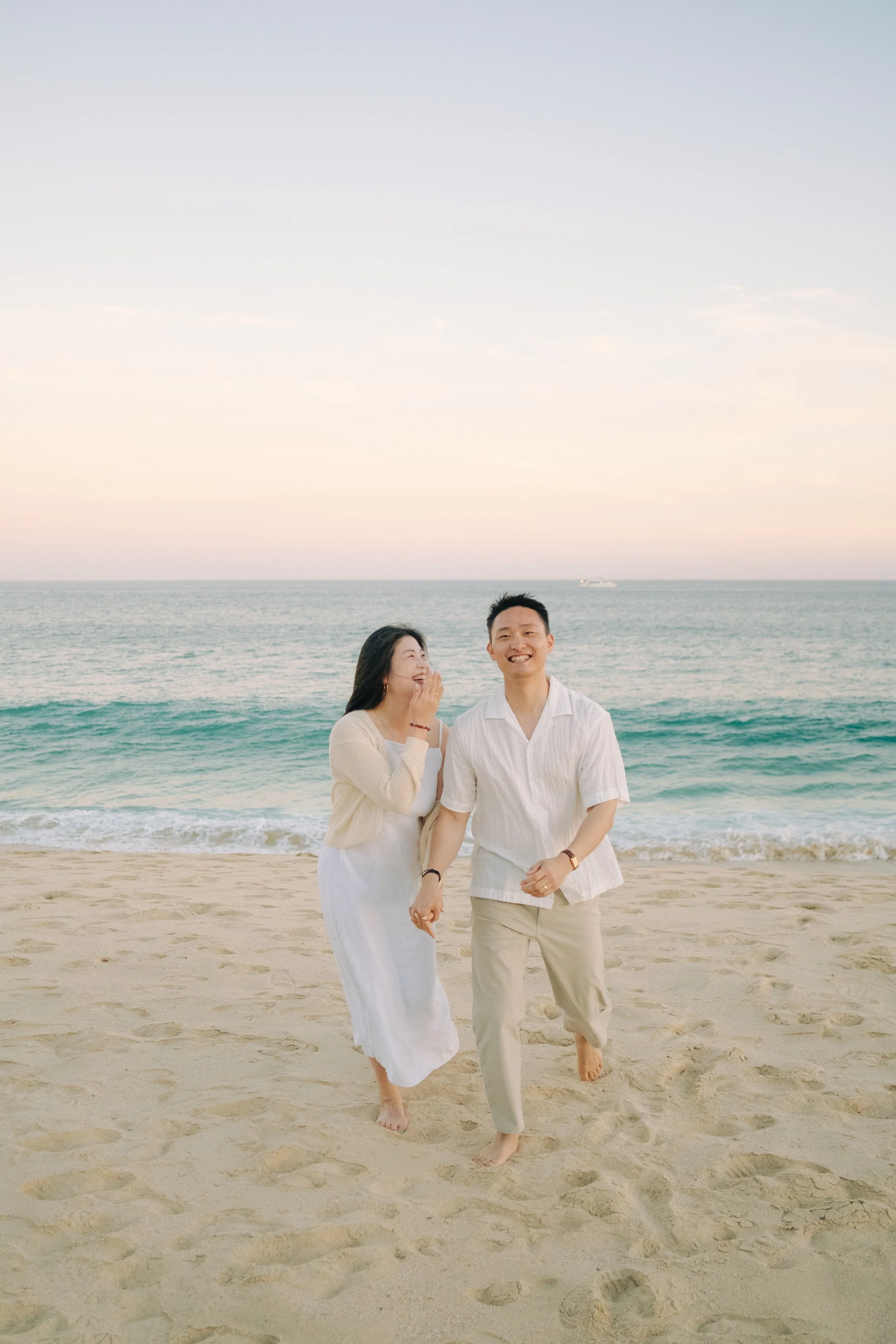 A couple walking on the beach holding hands, with the ocean and sunset in the background.