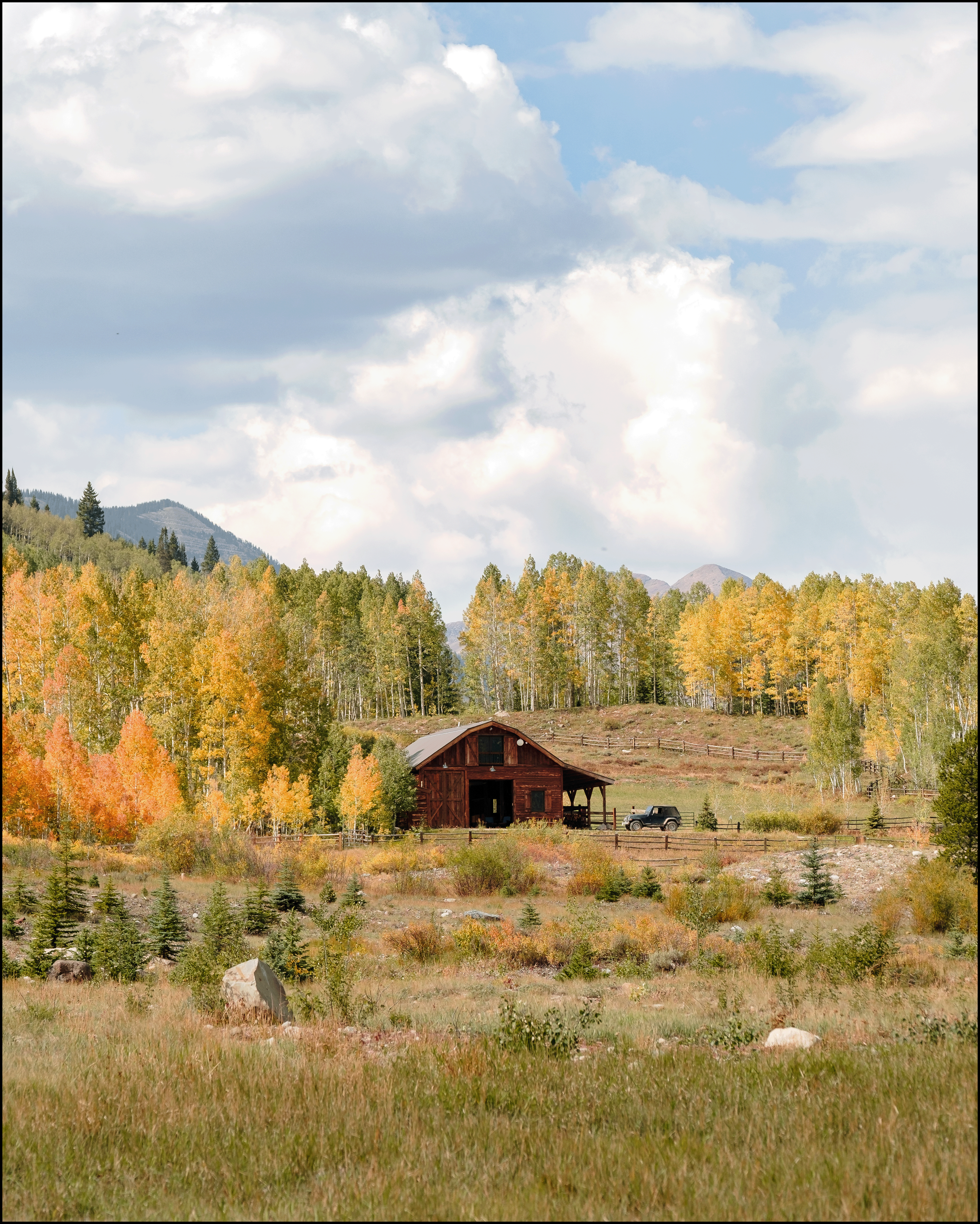 A wooden barn in a rural landscape with trees showing fall colors, a grassy field, and a partly cloudy sky.