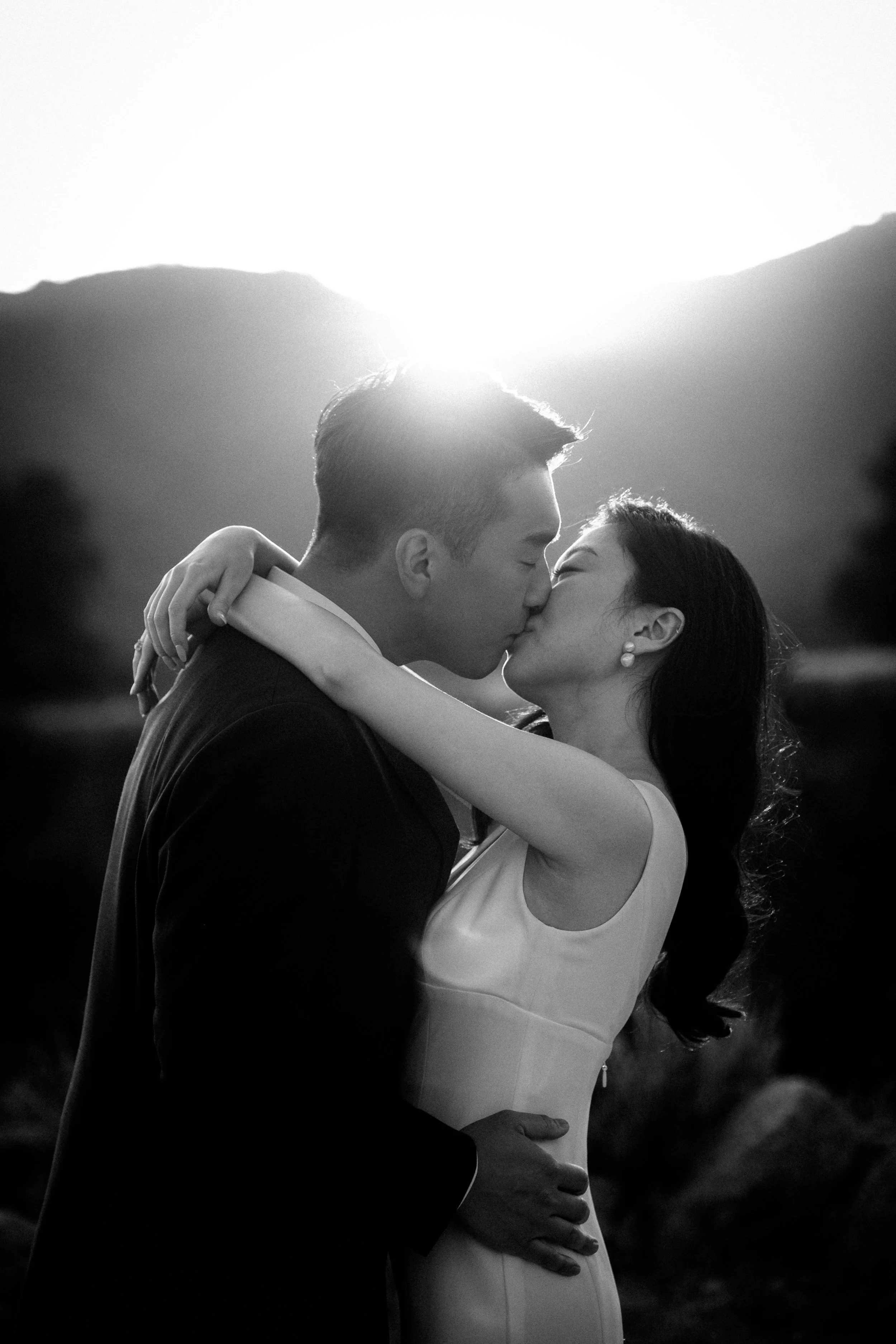 Black and white photo of a couple kissing outdoors with mountains in the background, sunlight shining behind them.