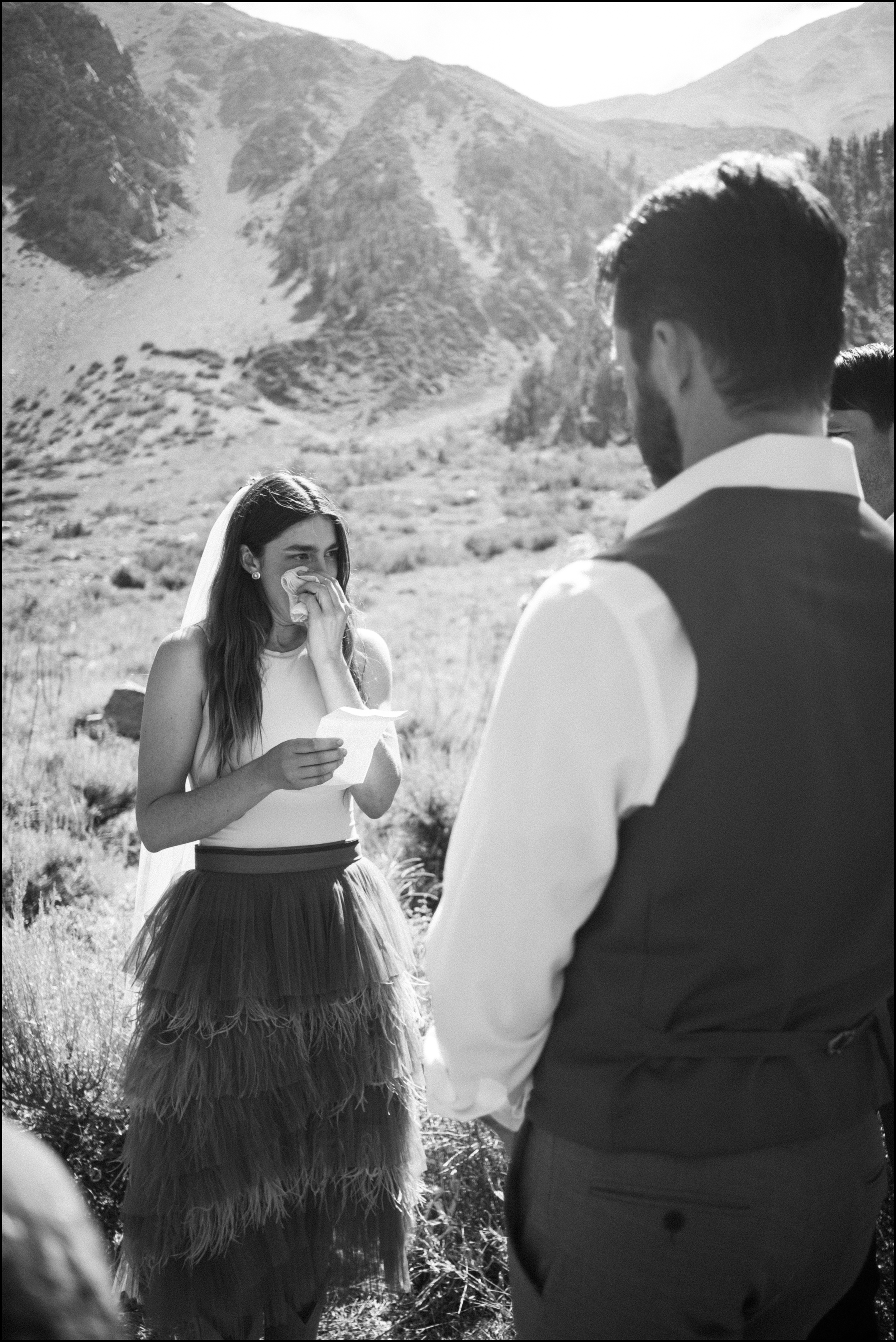 A woman wearing a wedding dress and veil is standing outdoors in a mountainous area, wiping away tears and holding a piece of paper, during her wedding ceremony with a man in formal attire.