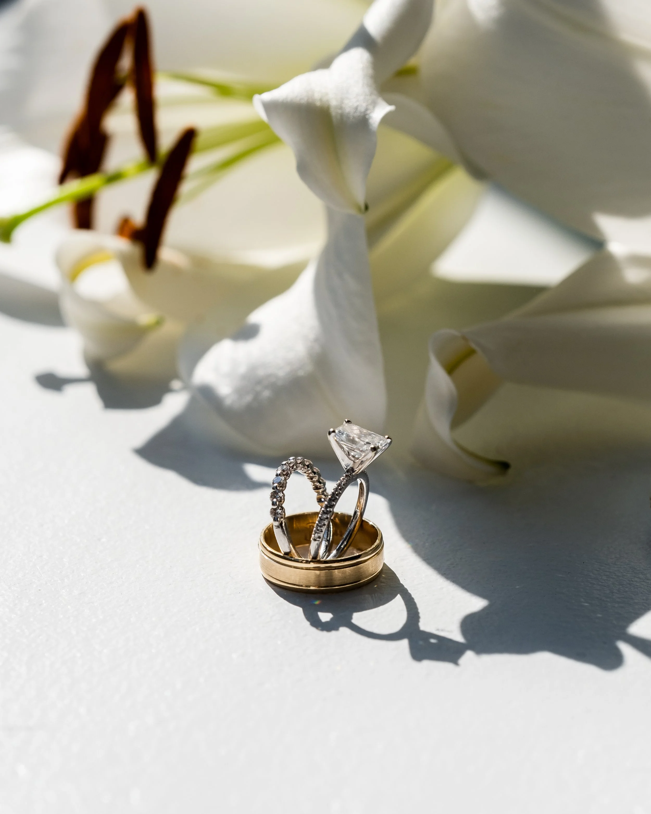 Close-up of three rings, including a diamond engagement ring, gold wedding band, and a diamond eternity band, placed on a white surface with white roses in the background.
