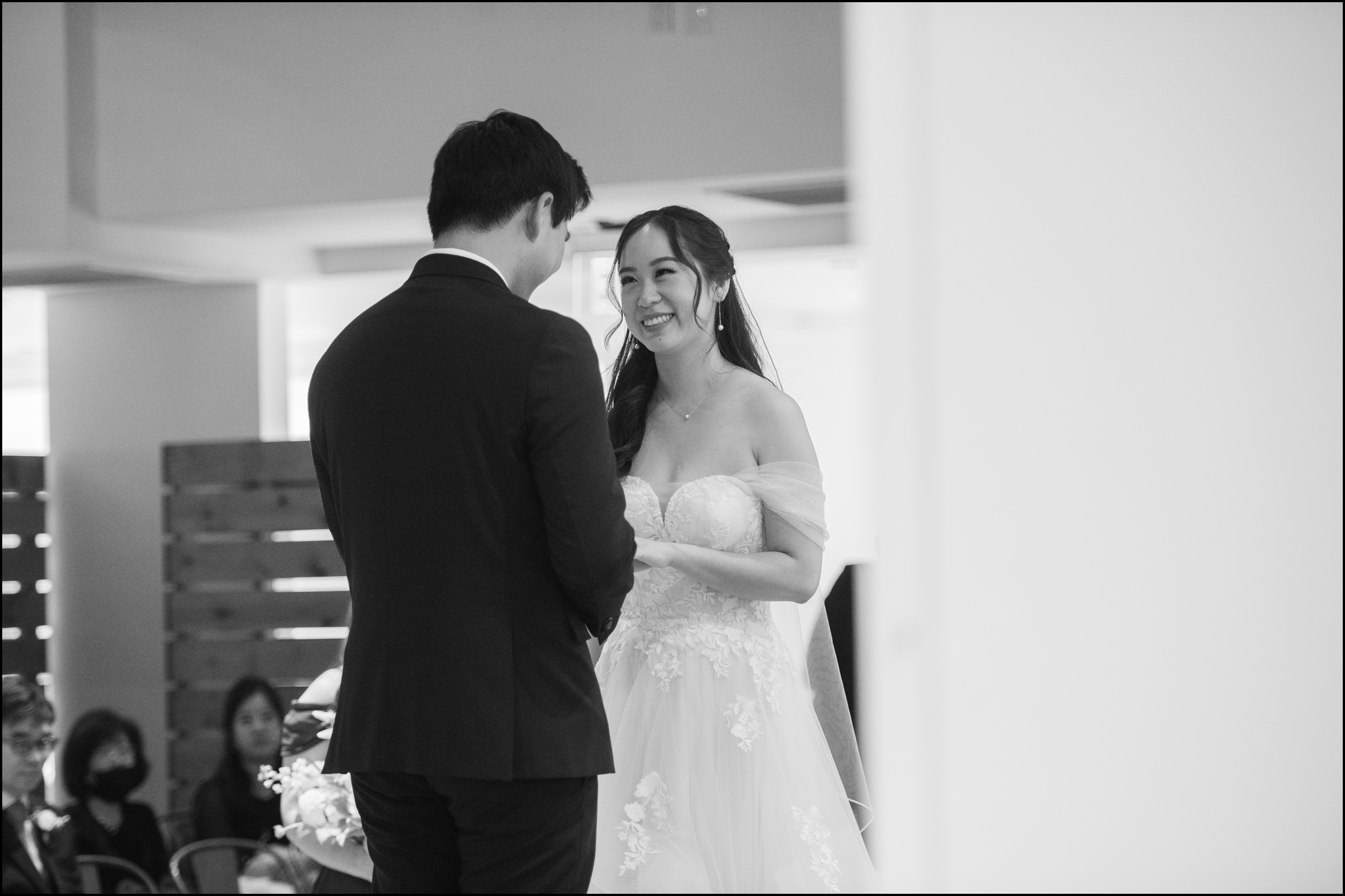 A couple exchanging vows during their wedding ceremony, with the bride smiling at the groom, in a modern indoor setting with guests watching.