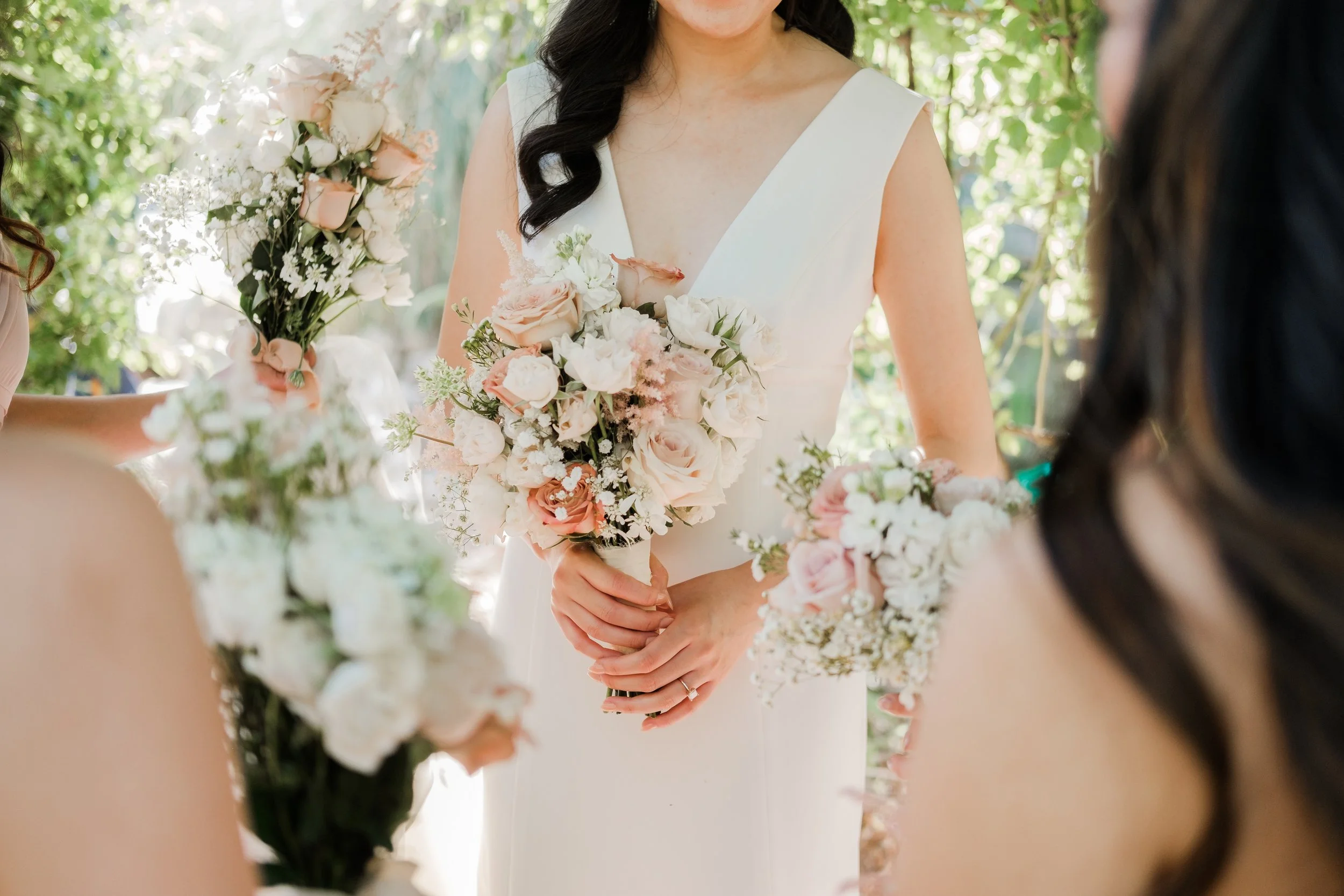 A bride holding a bouquet of pink and white flowers during a wedding ceremony outdoors, surrounded by bridesmaids in pastel dresses, with green trees in the background.