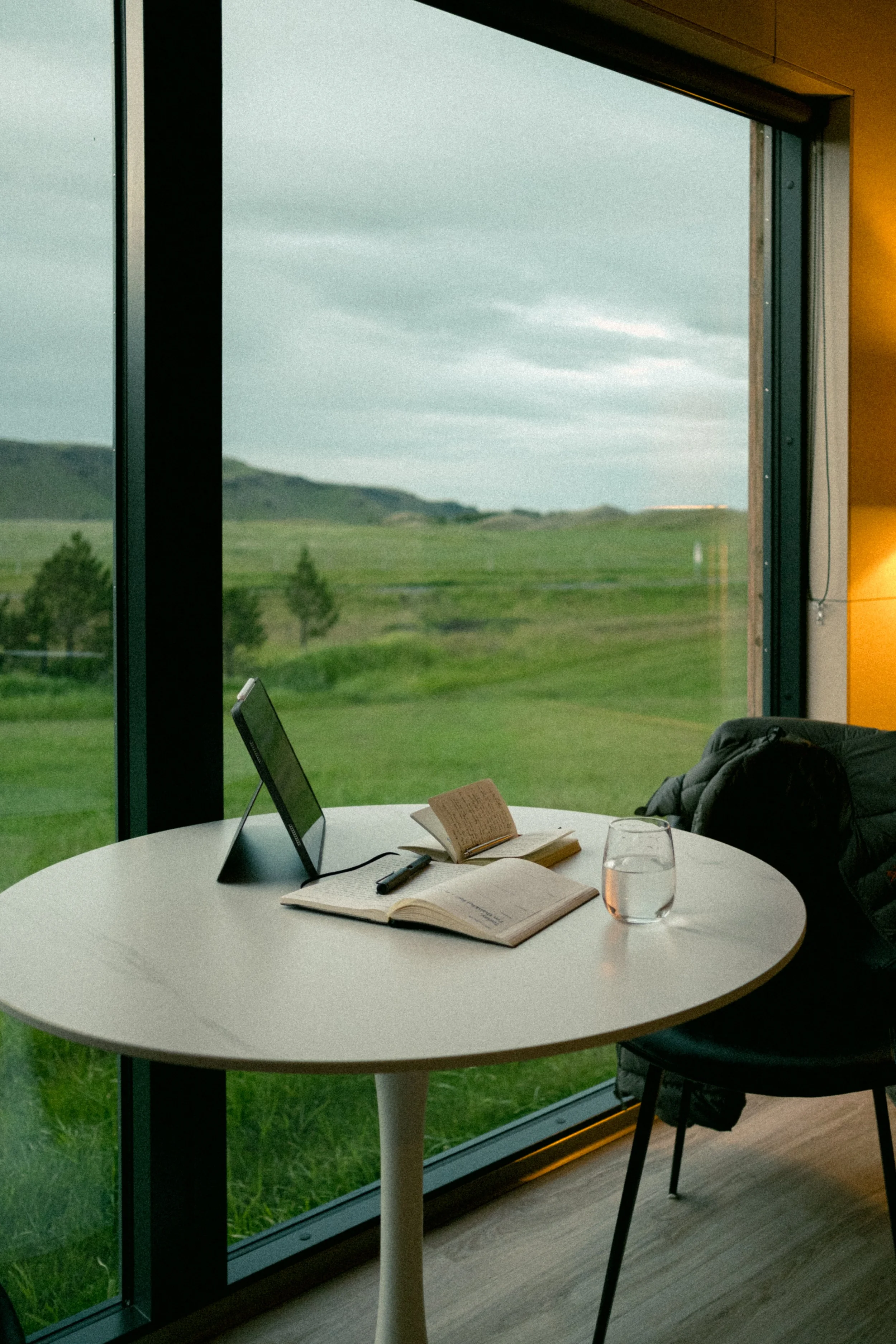 A round table with open notebooks, a pen, a digital device, and a glass of water near a large window overlooking green fields and hills.
