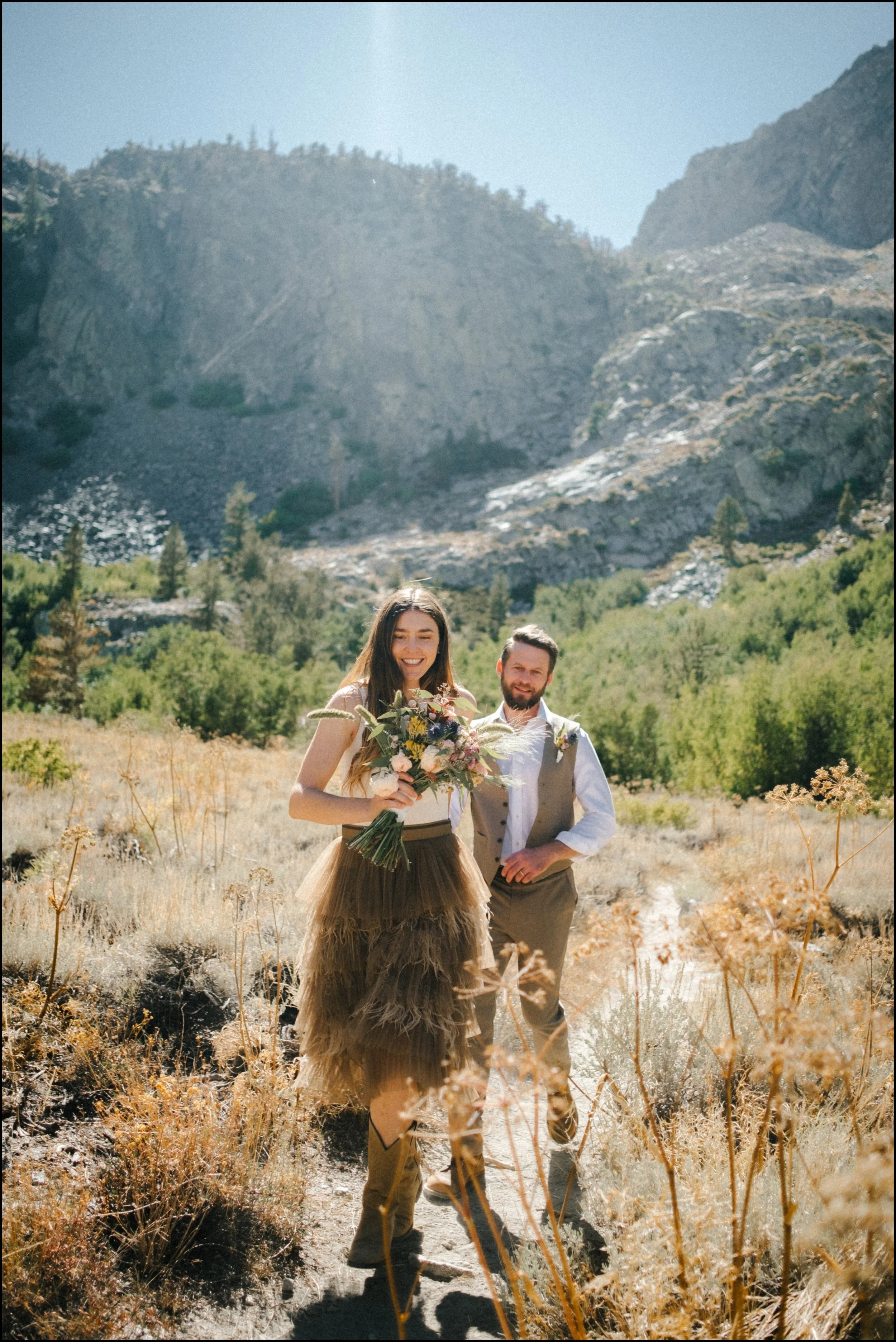 A woman and man outdoors on a trail in a mountainous area, with the woman holding a bouquet of flowers, both smiling.