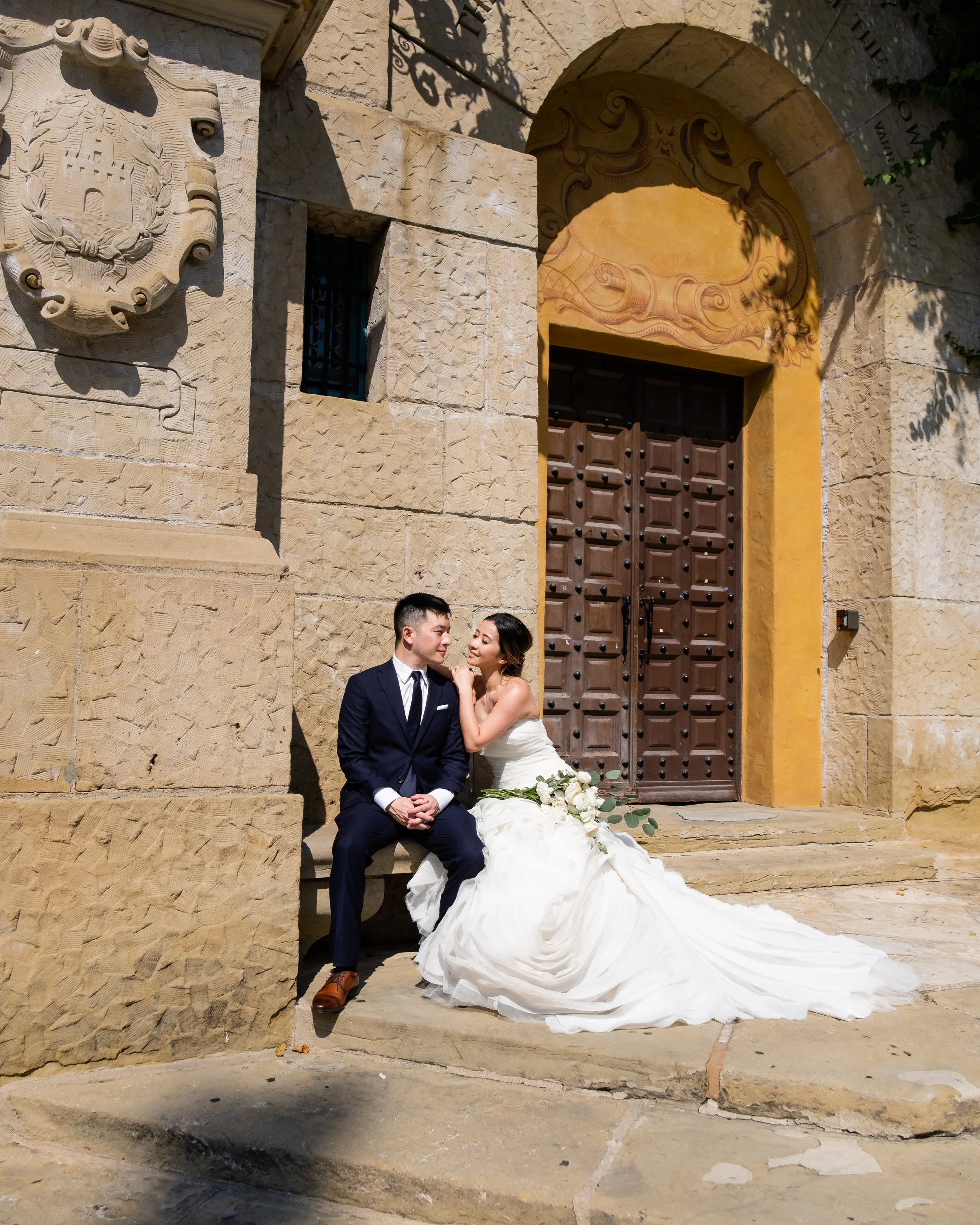 A bride and groom sit outside a stone building with a large wooden door. The bride is in a white wedding gown holding a bouquet, and the groom is in a dark suit. They are smiling and looking at each other.