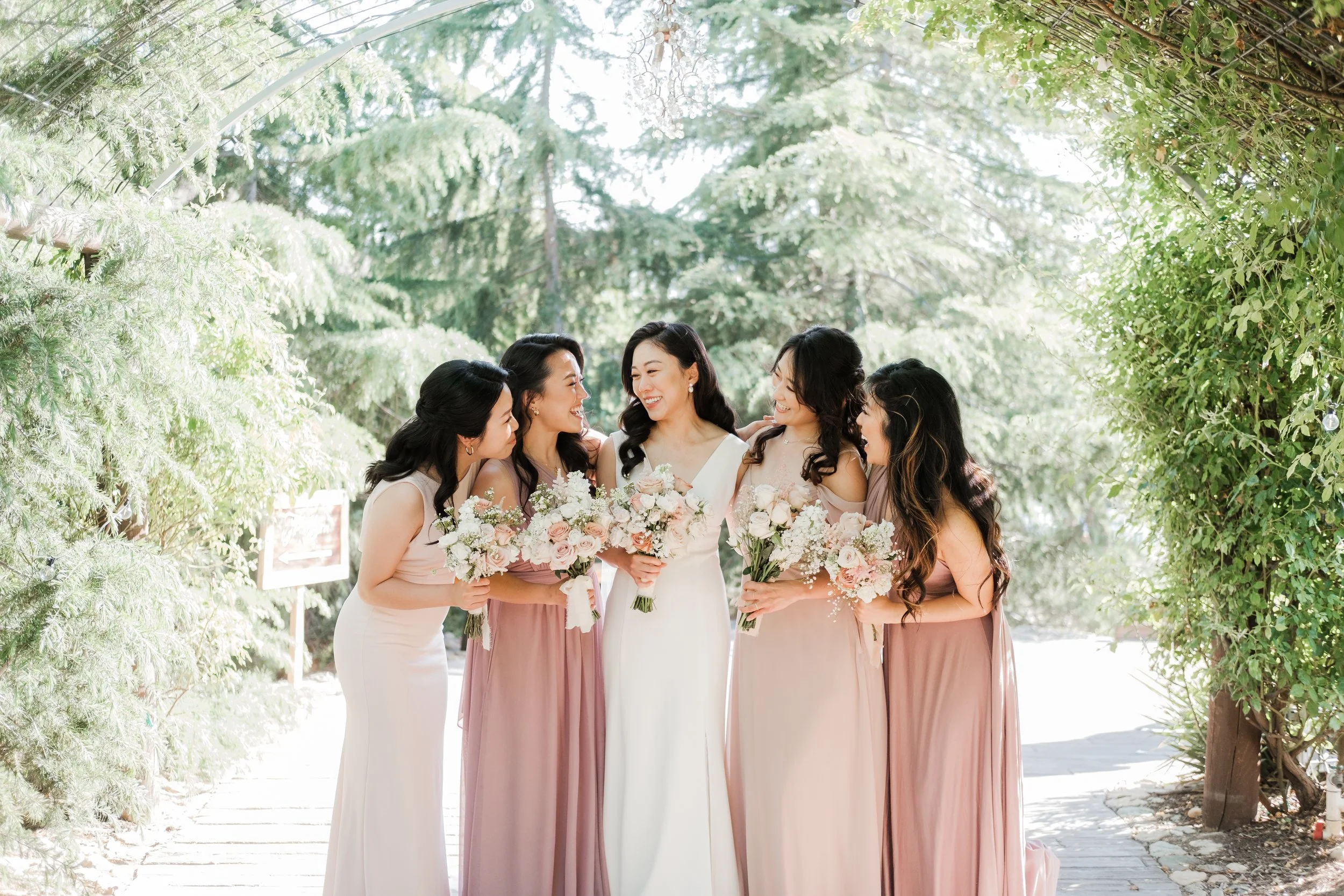 A bride and five bridesmaids standing outdoors surrounded by greenery, holding bouquets of flowers, smiling and enjoying each other's company.