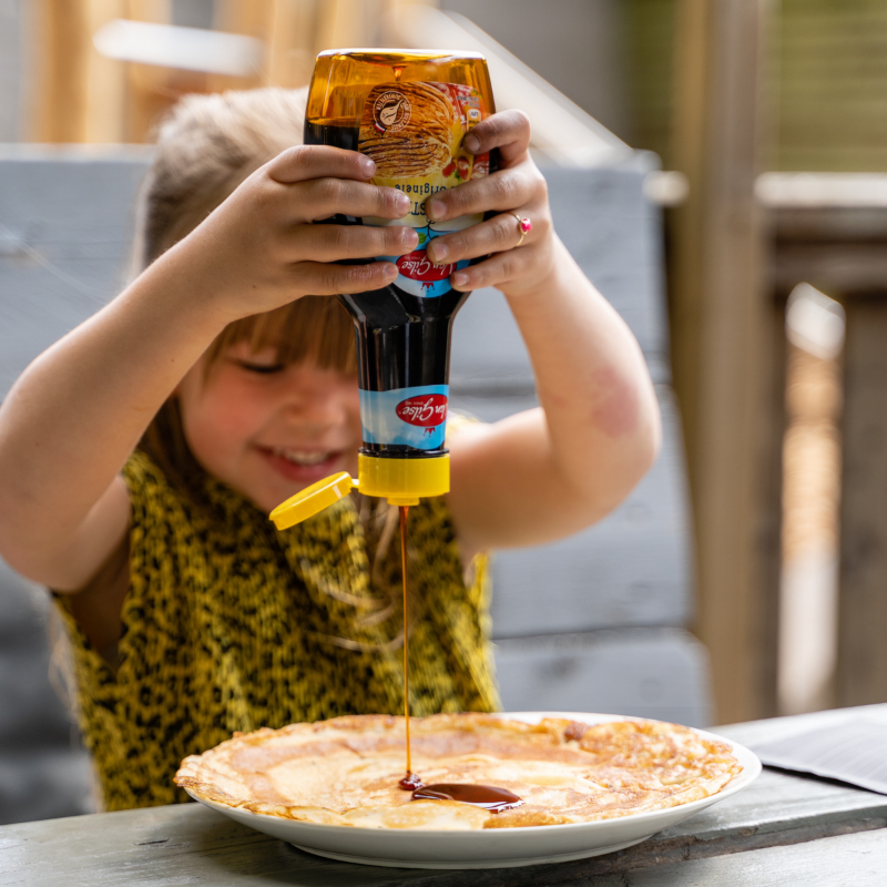A young girl with blonde hair and a yellow dress joyfully pouring mustard onto a pizza.