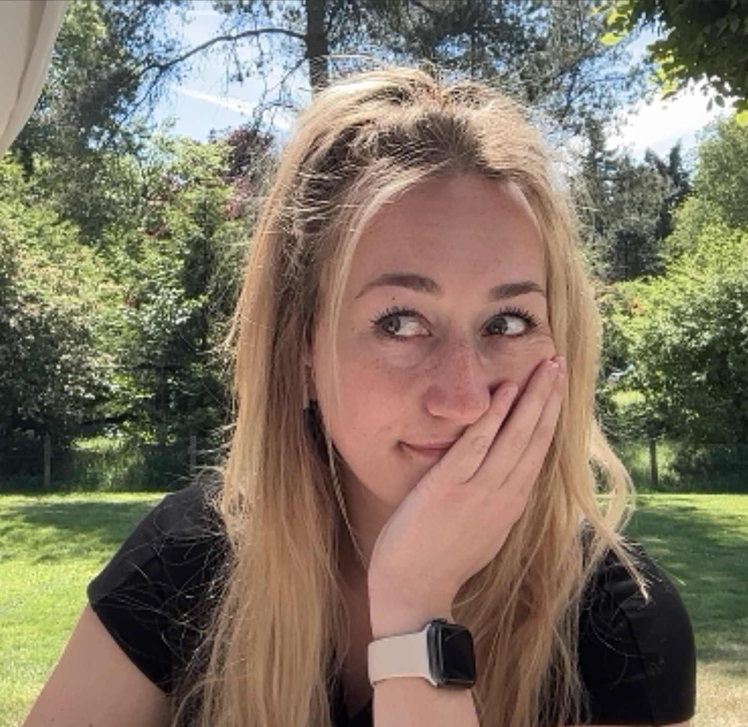 A young woman with blonde hair and freckles covering her mouth with her hand, outdoors on a sunny day with green trees and grass in the background.
