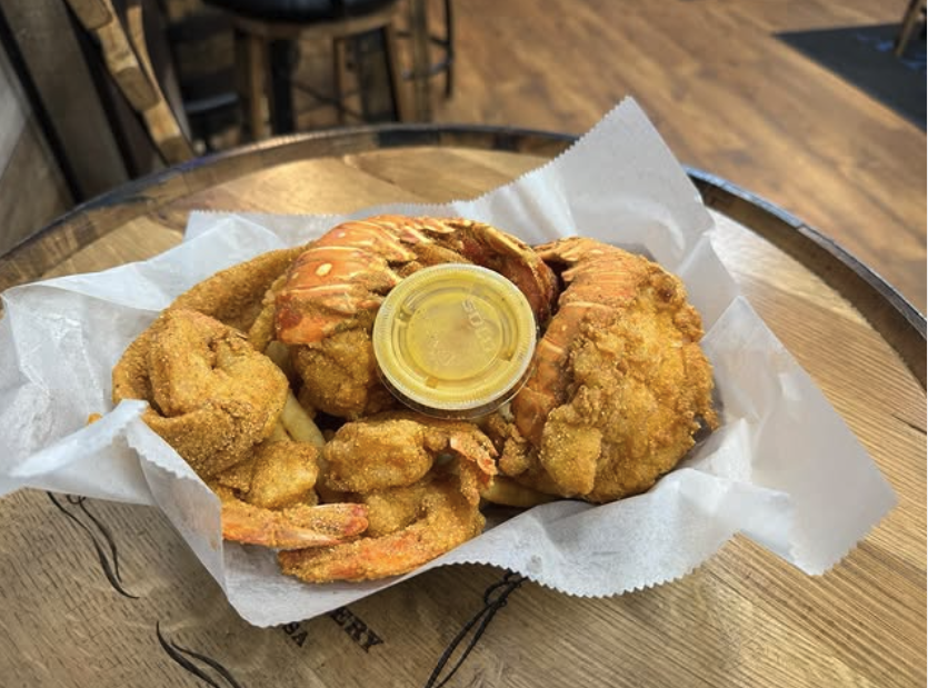 Fried seafood platter with shrimp and possibly fish, served with a cup of dipping sauce on a wooden tray lined with paper, in a restaurant setting.