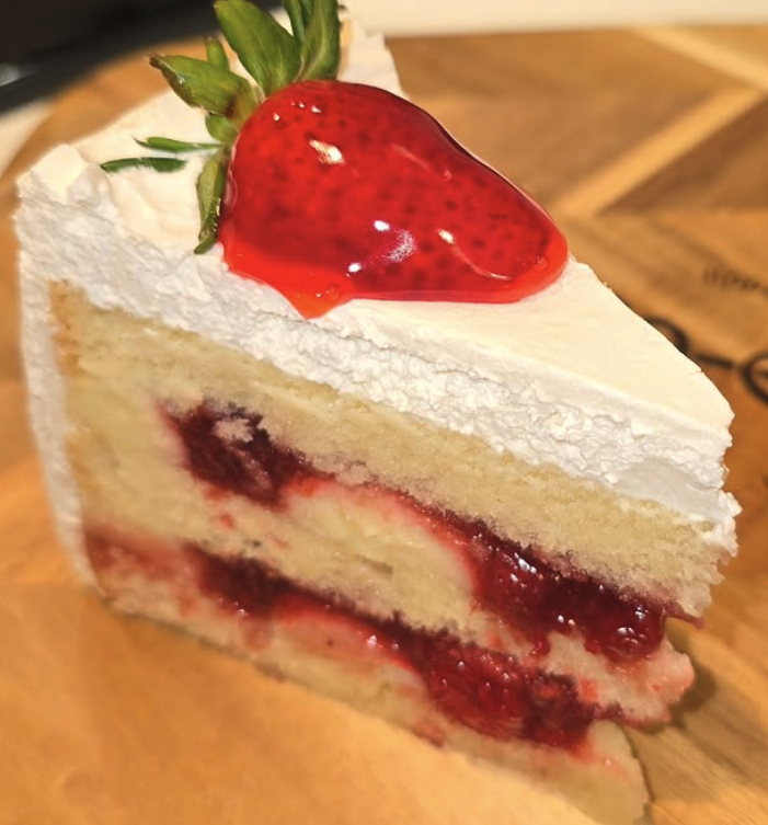 A slice of strawberry shortcake with white frosting, layers of sponge cake, and strawberry filling, topped with a fresh strawberry.