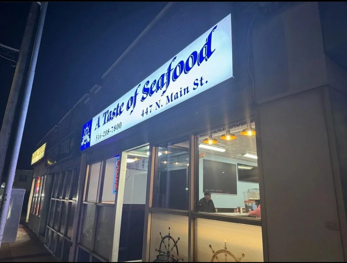 Nighttime exterior view of a seafood restaurant called 'A Taste of Seafood' with signage displaying the address 447 N. Main St. and phone number 516-208-7800, with visible interior through windows showing a person inside.