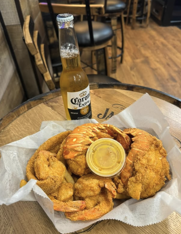 Fried shrimp with two breaded onion rings and a small container of dipping sauce on white paper lining on a wooden table with a Corona Extra beer bottle in the background.