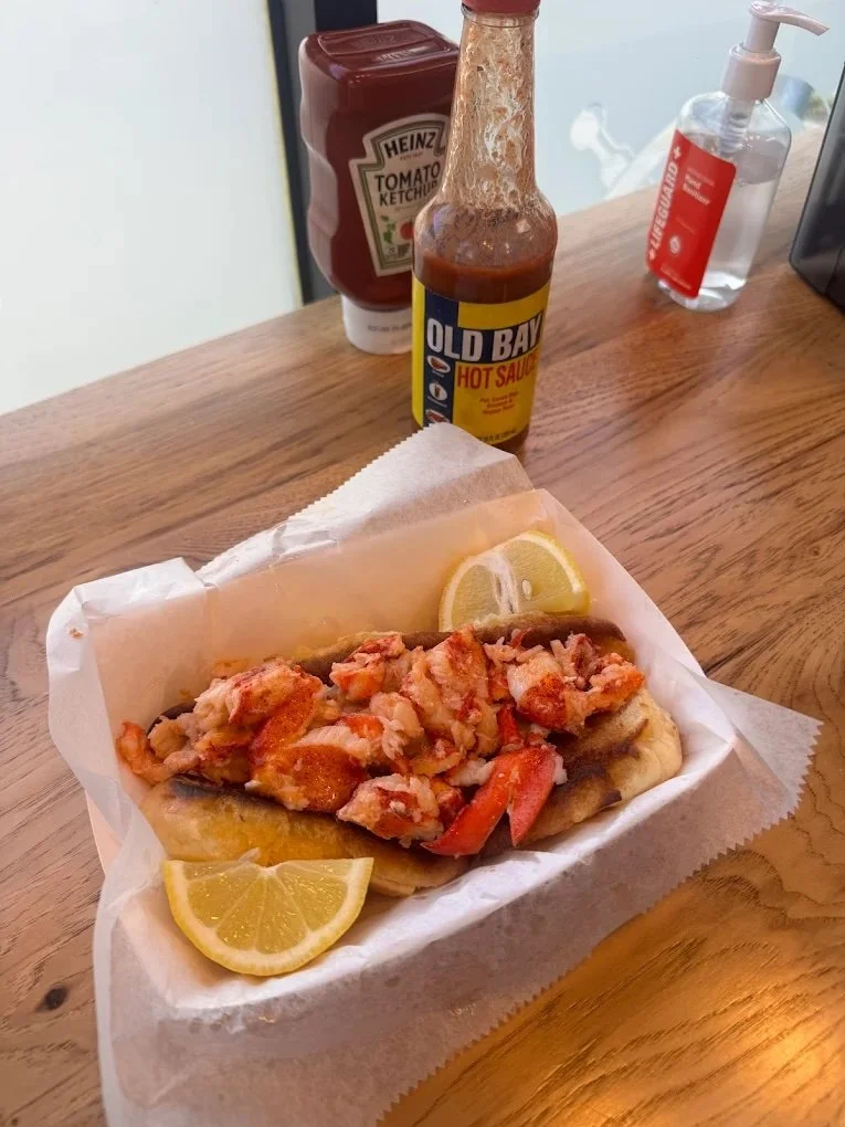 Lobster roll with lemon slices on a paper-lined basket, condiments including ketchup, hot sauce, and hand sanitizer on a wooden table.