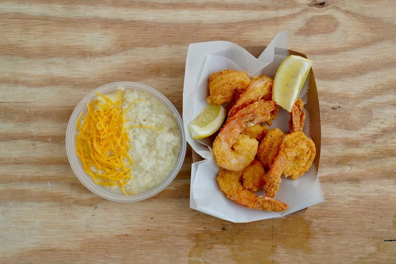 Fried shrimp with lemon wedges served on a paper-lined tray, alongside a cup of cheesy grits with shredded cheddar cheese, on a wooden surface.