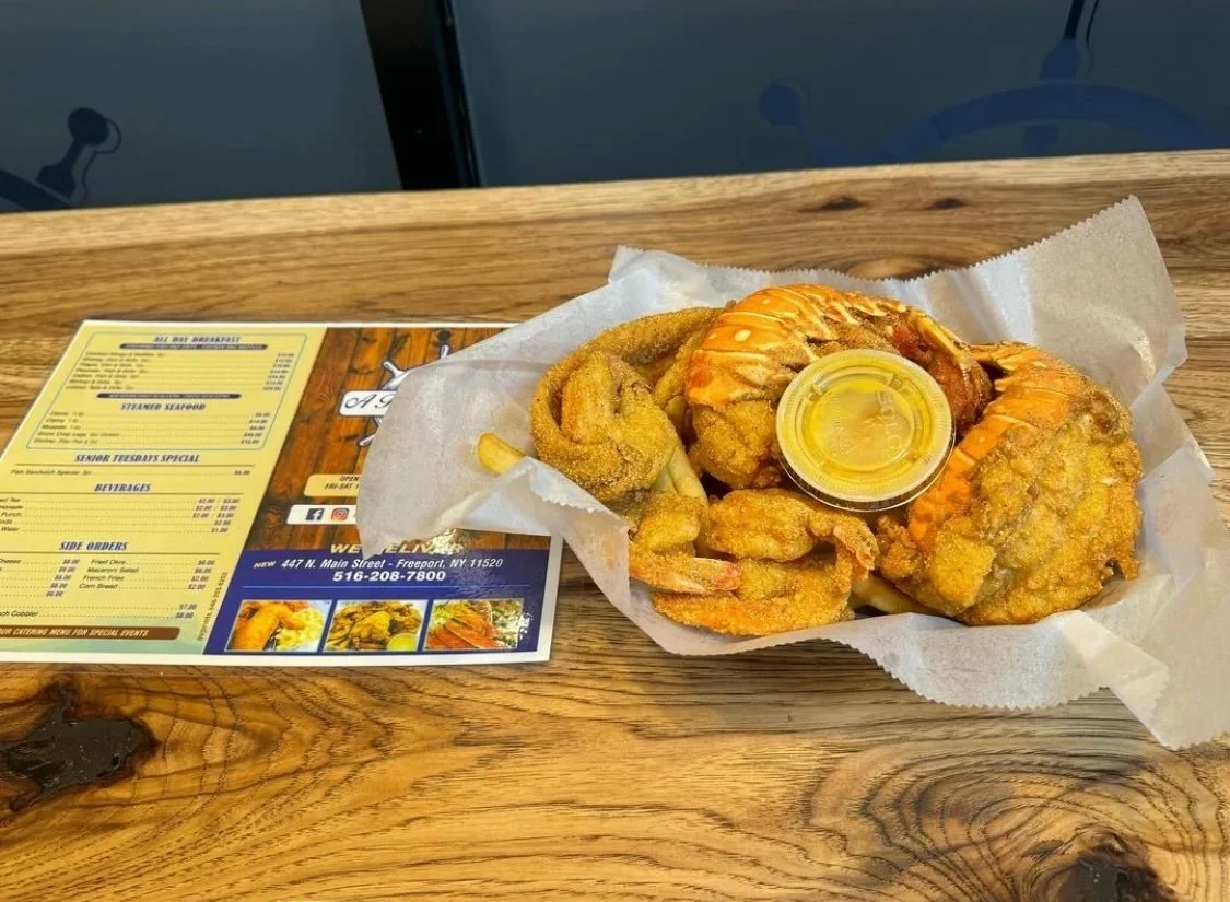 Fried seafood platter with shrimp and fish, served with a small container of yellow sauce on white parchment paper on a wooden table, with a restaurant menu nearby.
