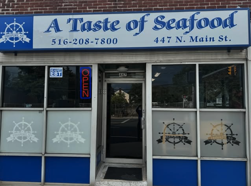 Front view of a seafood restaurant called 'A Taste of Seafood' with a blue and white sign, phone number 516-208-7800, address 447 N. Main St., and a neon 'Open' sign. The restaurant has large windows with the logo featuring a ship's wheel and the tex