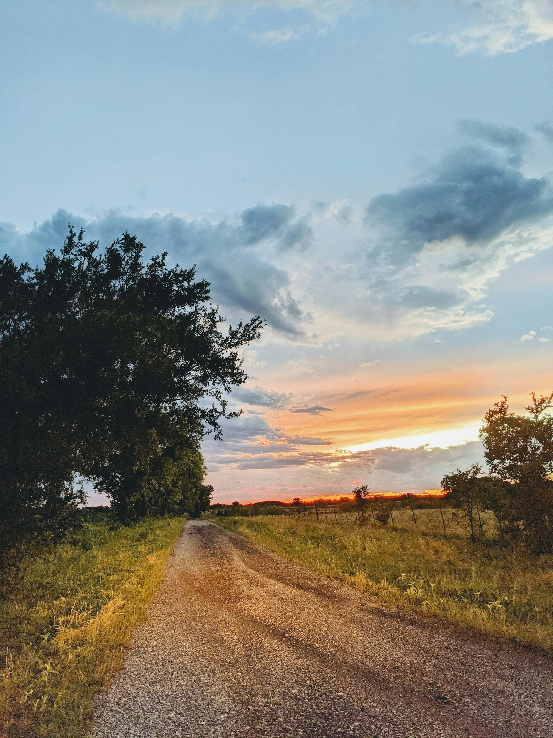 A dirt country road with grass on both sides, lined with trees and a fence, under a colorful sunset sky with clouds.