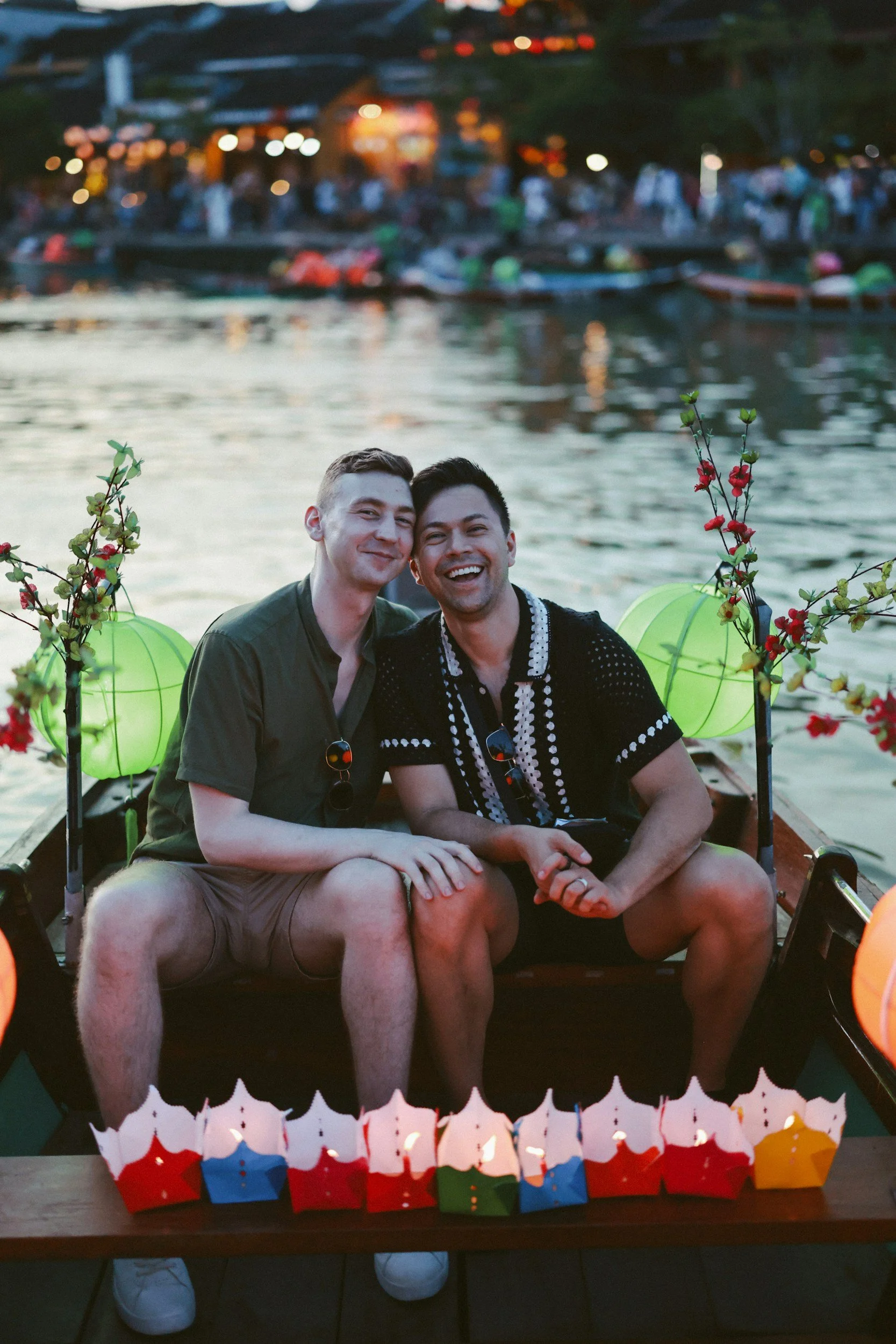 Two men sitting on a decorated boat near a body of water during evening. The background shows a crowd and boats on the water, with lighting from lanterns and a lively atmosphere.