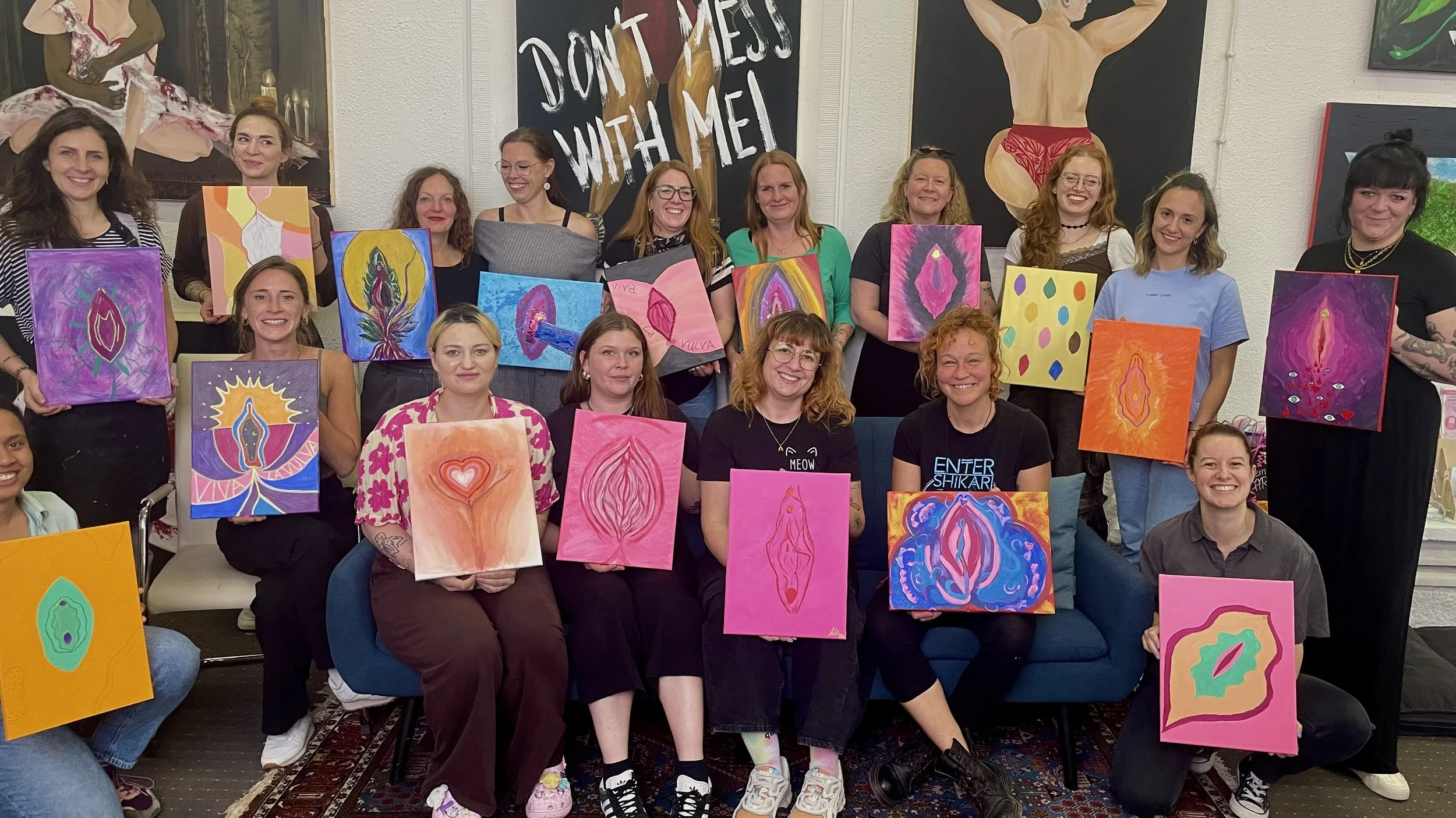 Gruppe von Menschen, die bei einem Kunstkurs ihre Gemälde präsentieren, mit einem Wandbild mit dem Text "DON'T MESS WITH ME" im Hintergrund.