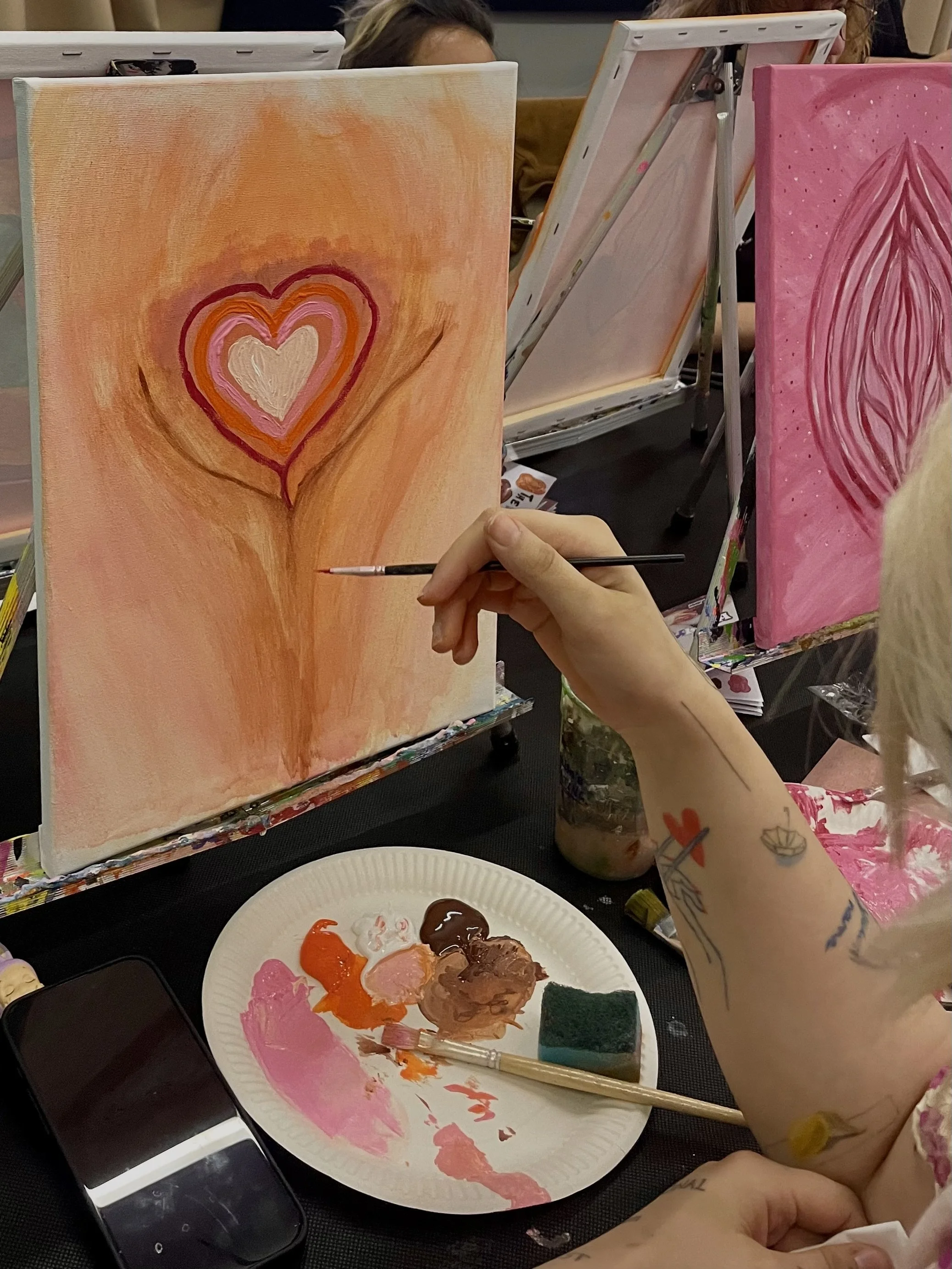 Malerin beim Malen einer Vulva mit Herz auf einer Leinwand mit verschiedenen Pink-, Orange- und Weißtönen.
