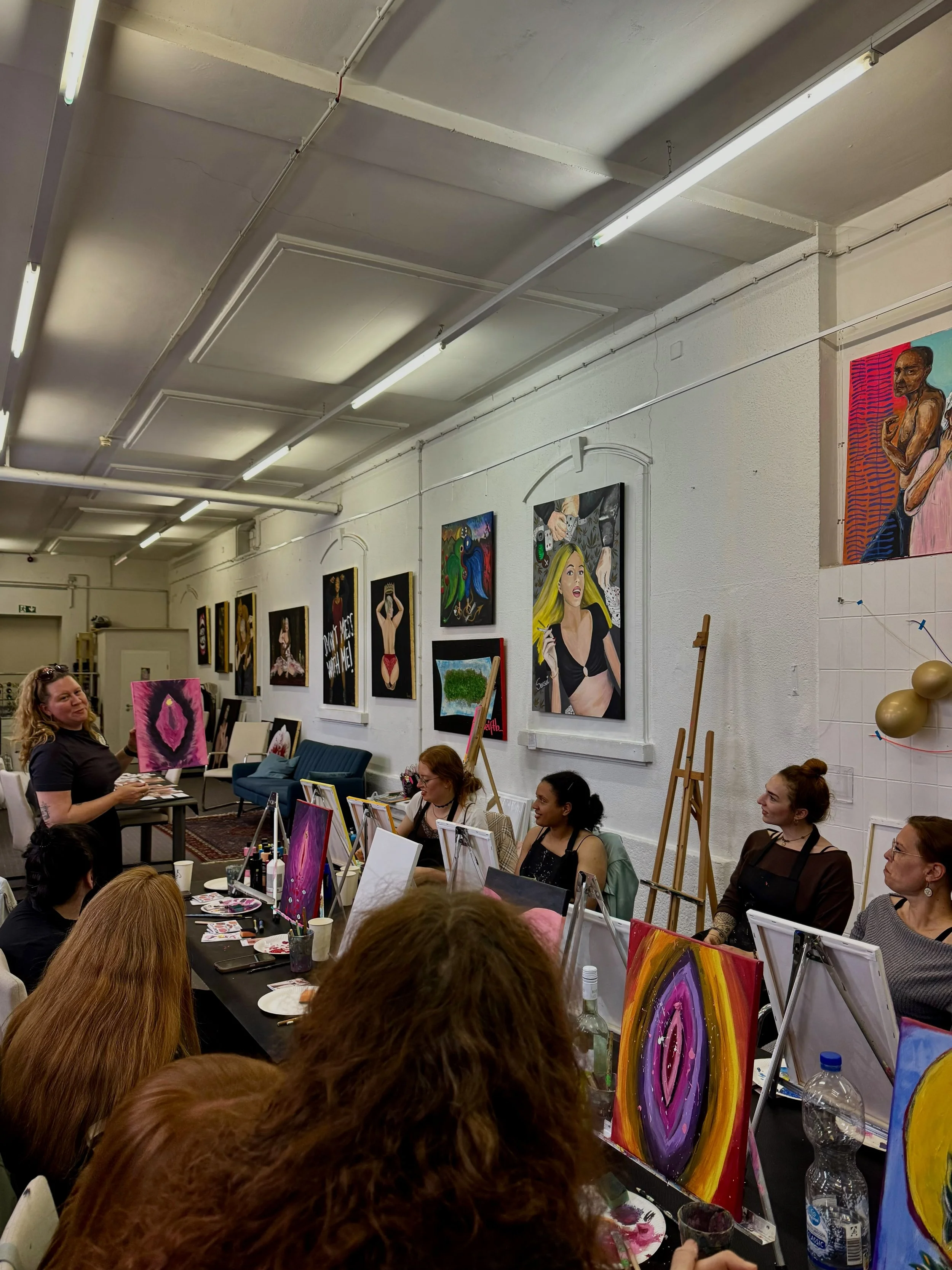 Menschen, die in einem Raum bei einem Mal-Workshop ihre Kunstwerke malen, mit bunten Gemälden an der Wand im Hintergrund.