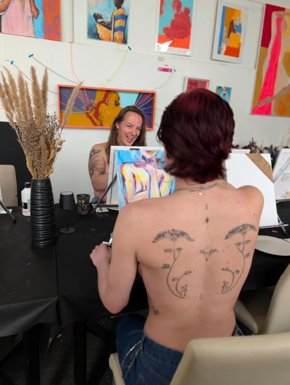 Zwei Personen beim Malen in einem Kunststudio, die eine lacht. Eine sitzt mit Rückenansicht, trägt keine Oberbekleidung, hat Tattoos auf dem Rücken, die andere sitzt vor einem Bildschirm und malt.