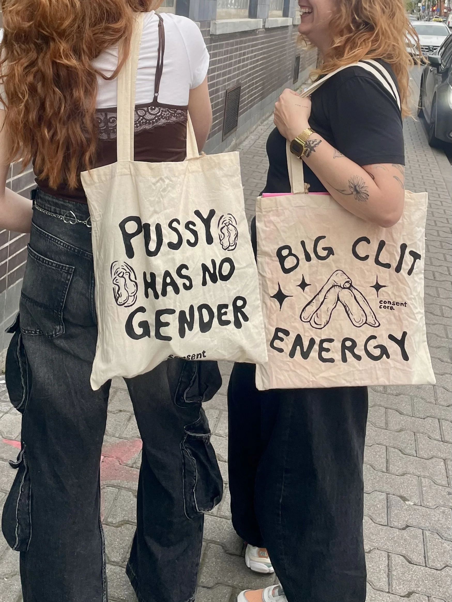 Zwei Menschen stehen sich auf der Straße gegenüber, tragen Tragetaschen mit feministischen Botschaften, lachen und unterhalten sich.