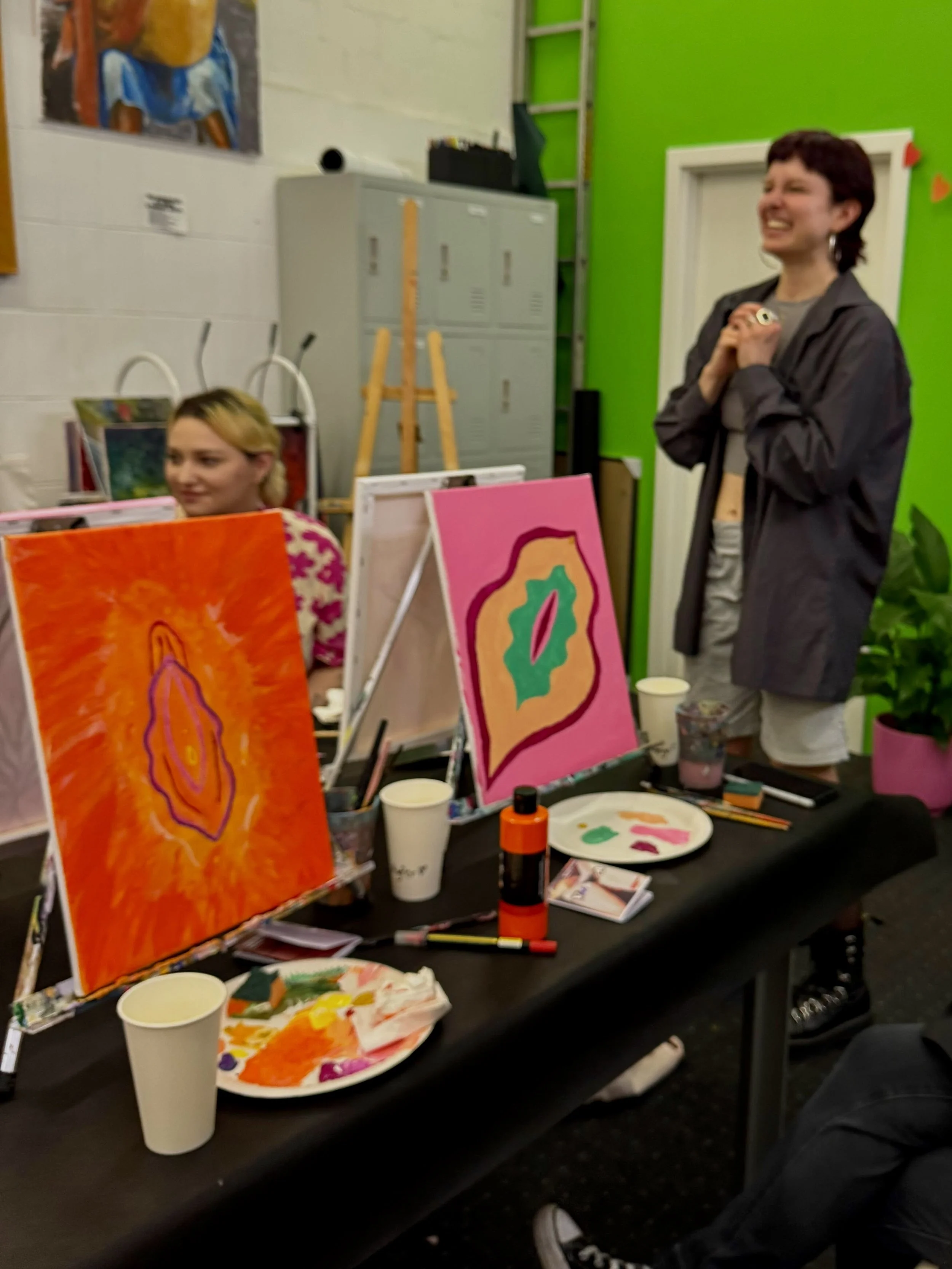 Zwei Menschen beim Malen von abstrakten Kunstwerken in einem Raum mit grüner Wand und Kunst auf der Wand. Tische mit Farbpaletten, Malutensilien und Papieren.