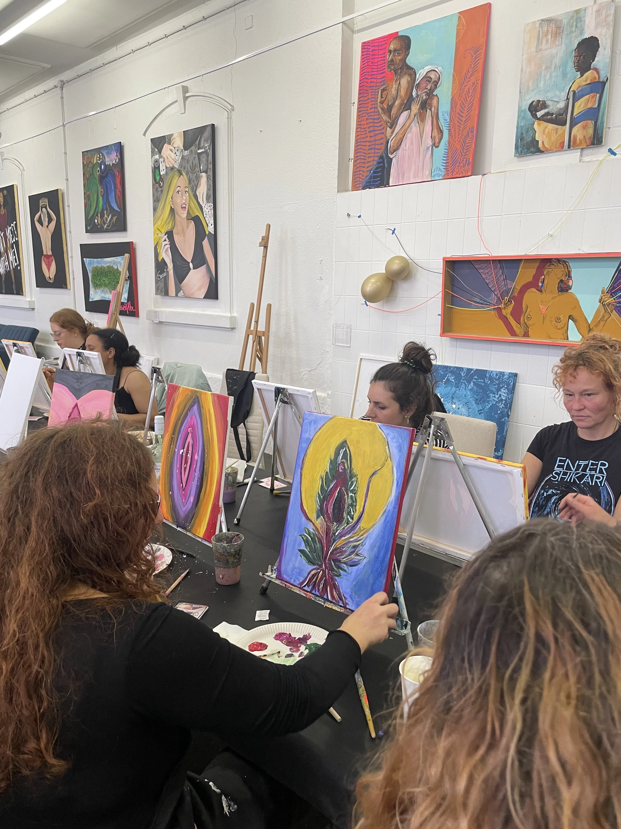 Menschen beim Malen in einem Kunstkurs, mit verschiedenen Farben und Leinwänden, in einem gut beleuchteten Raum mit weißen Wänden und mehreren aufhängenden Kunstwerken.