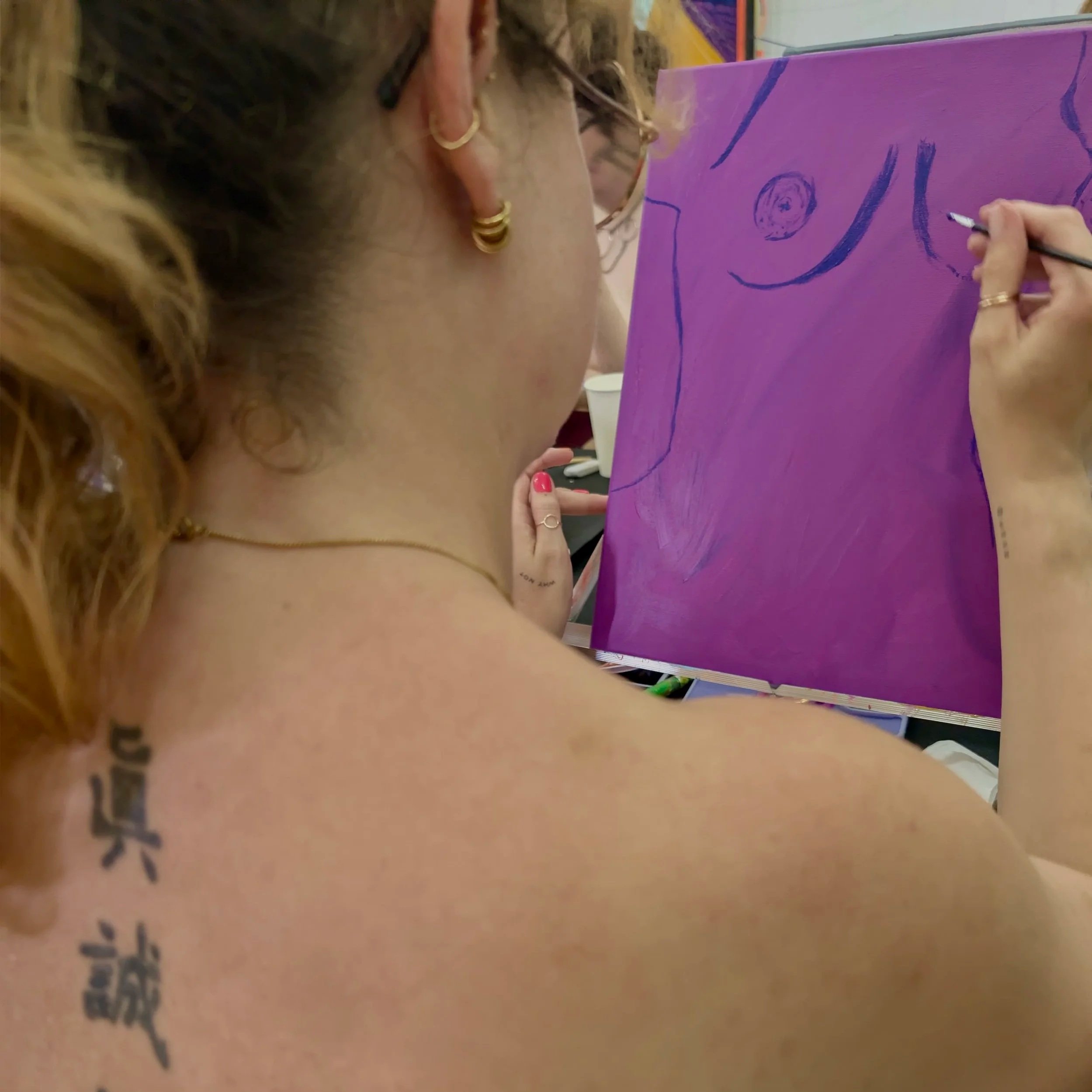 Eine Person mit lockigem, blondem Haar trägt mehrere goldene Ringe und eine goldene Kette. Die Person zeichnet auf eine Leinwand.