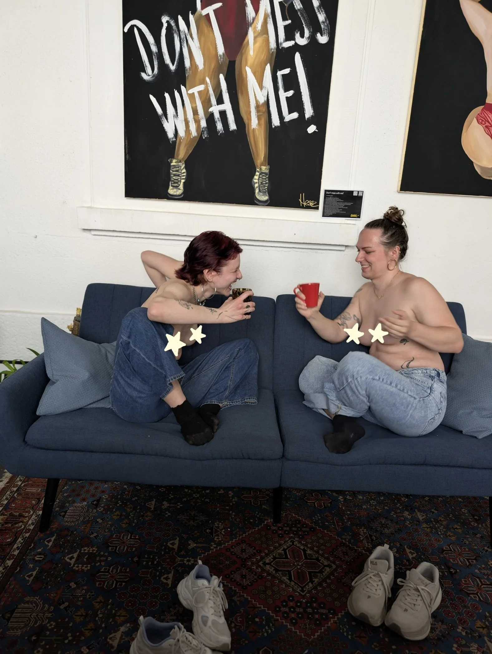 Zwei Personen sitzen auf einem blauen Sofa, lachen und trinken aus Tassen. Im Vordergrund sind weiße Turnschuhe auf einem bunten Teppich zu sehen. Die Personen sind oberkörperfrei, haben Tattoos und tragen Ohrringe.