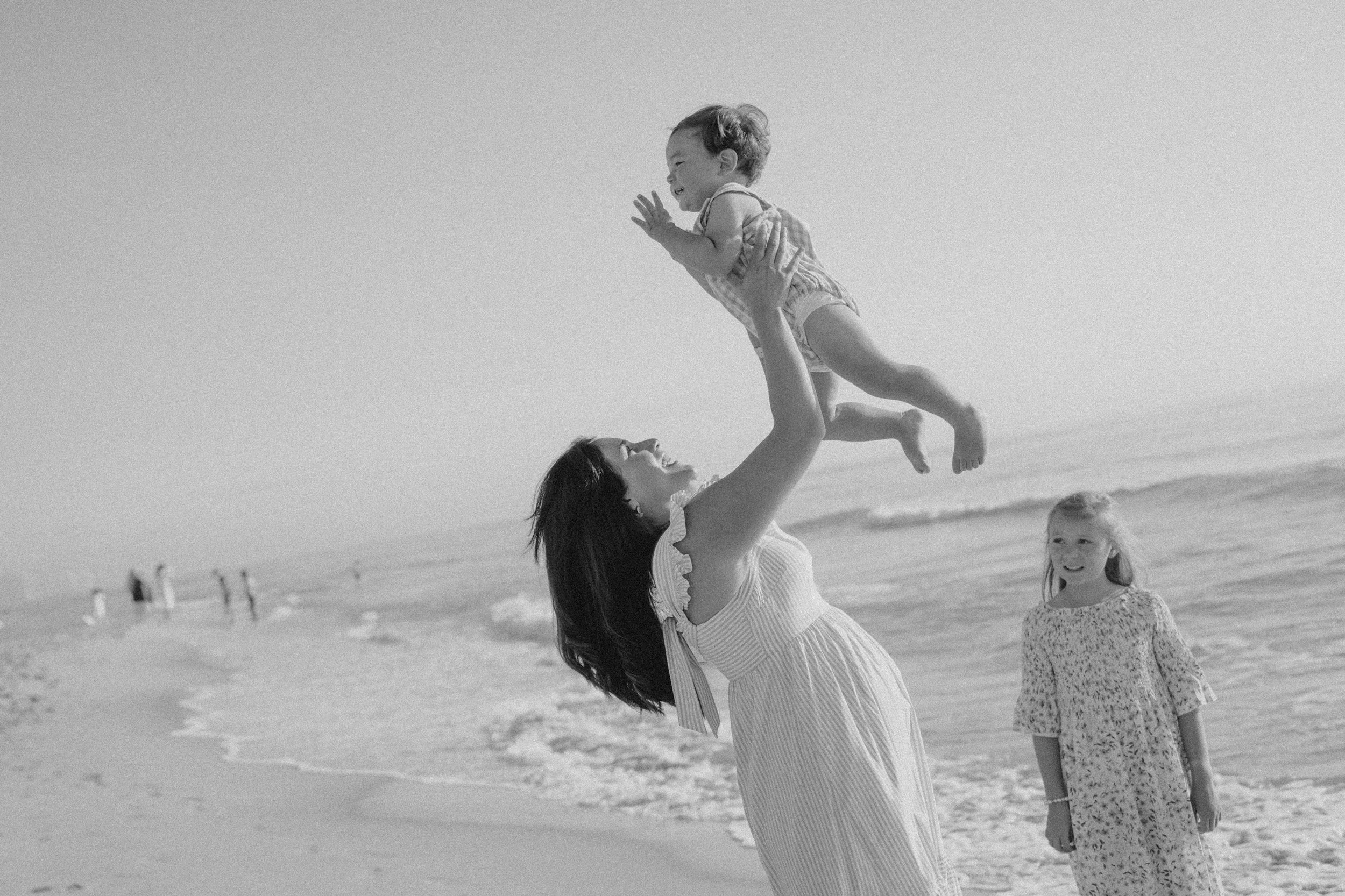 A mother, who used The Birth Atelier's daytime postpartum doula services in New Jersey, throws her child in the air at the beach.