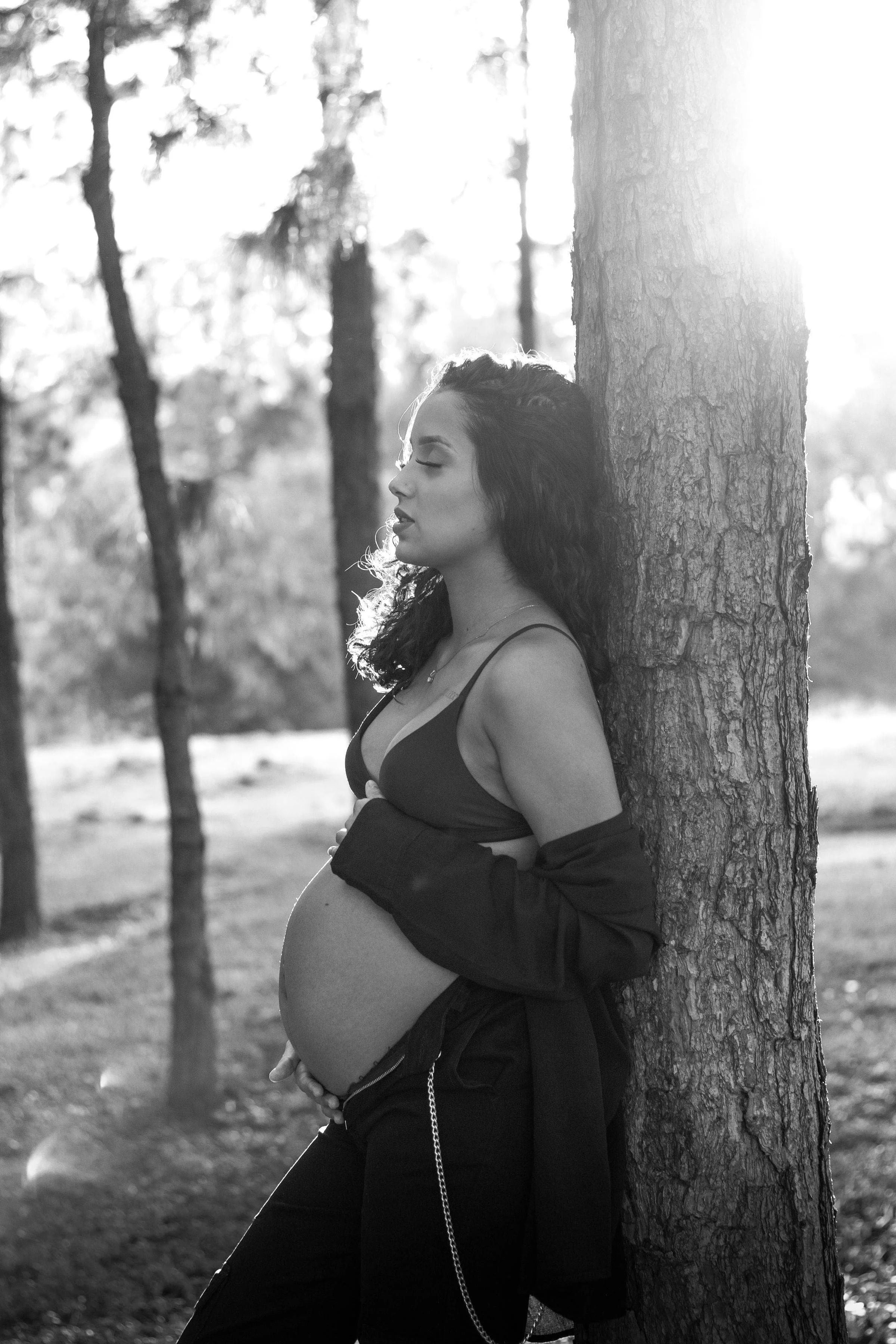 A pregnant mother holds her stomach while leaning against a tree in New Jersey, feeling at peace with her birth doula support services from The Birth Atelier.