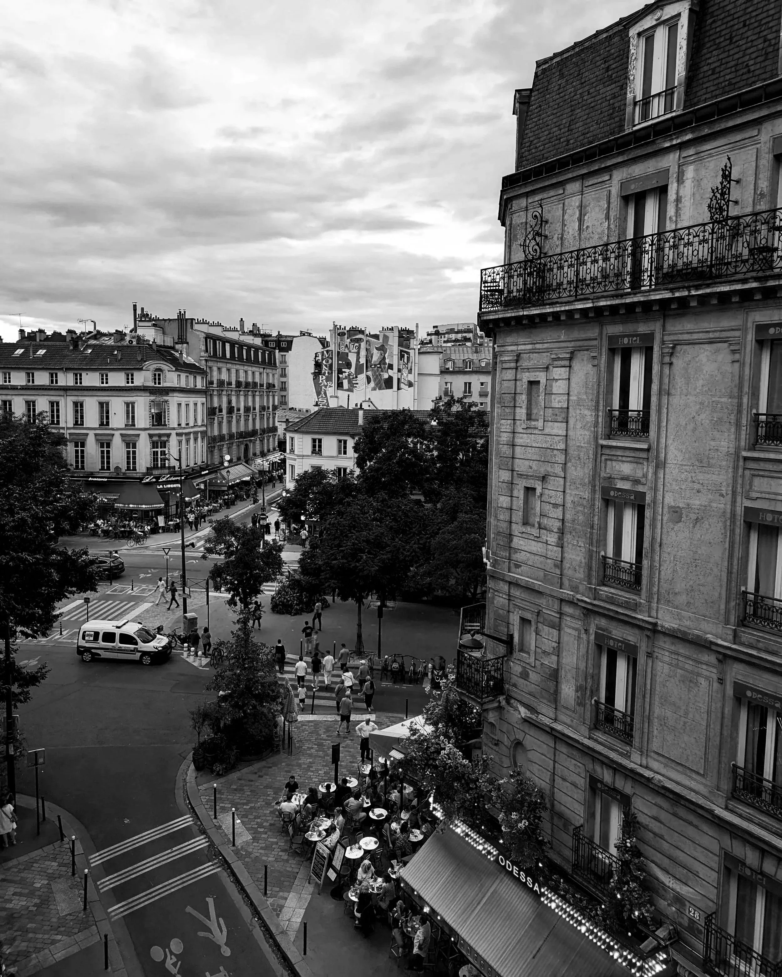 Black and white photo of a busy Parisian street corner with multiple buildings, cafes, trees, and pedestrians, including outdoor seating and a small van.