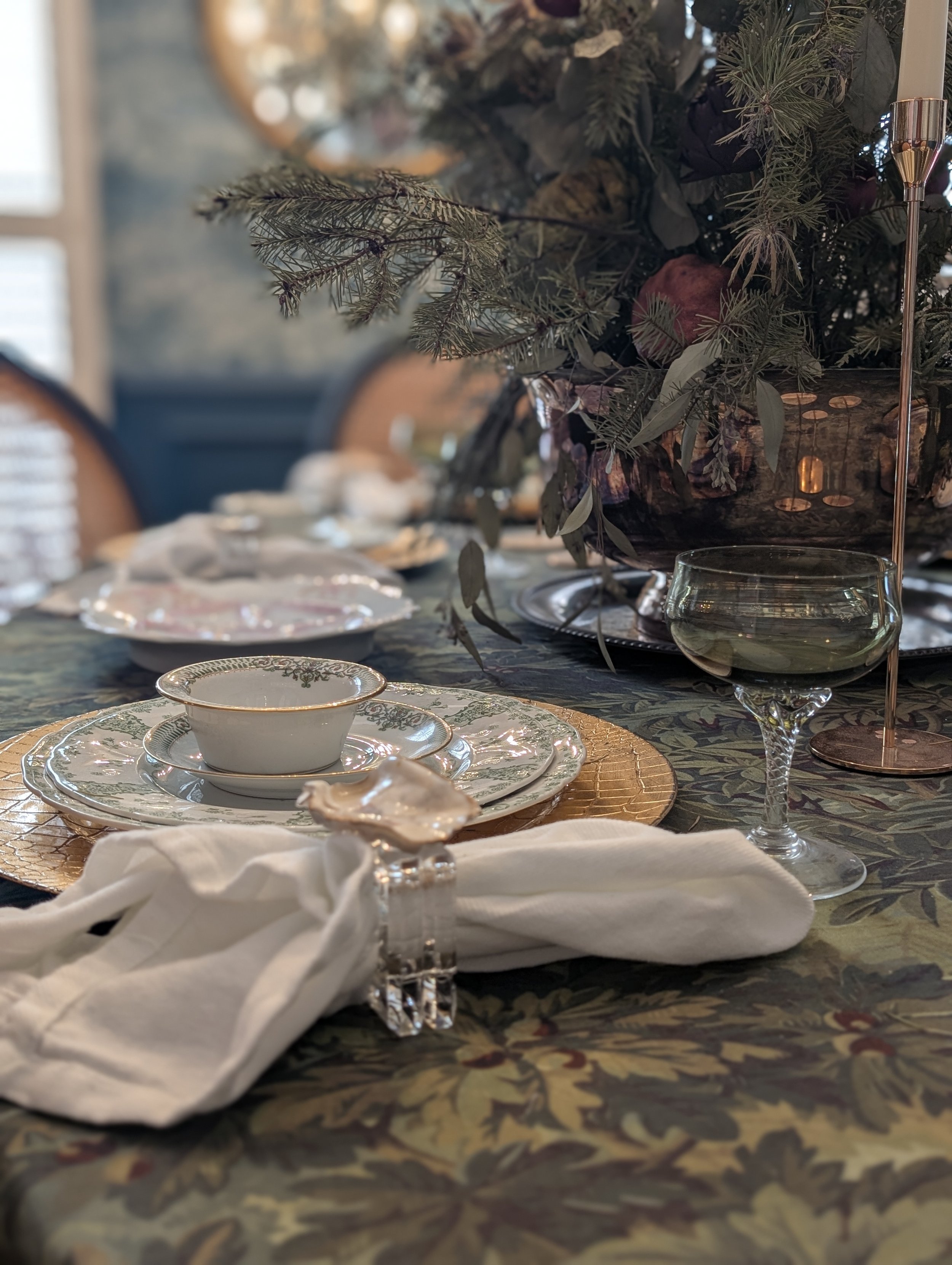 Elegant dinner table with crystal glassware, fine china, and white cloth napkin. In the background, a large floral arrangement with greenery and dark flowers. The tablecloth has a dark floral pattern, and there is a gold placemat under the table setting.