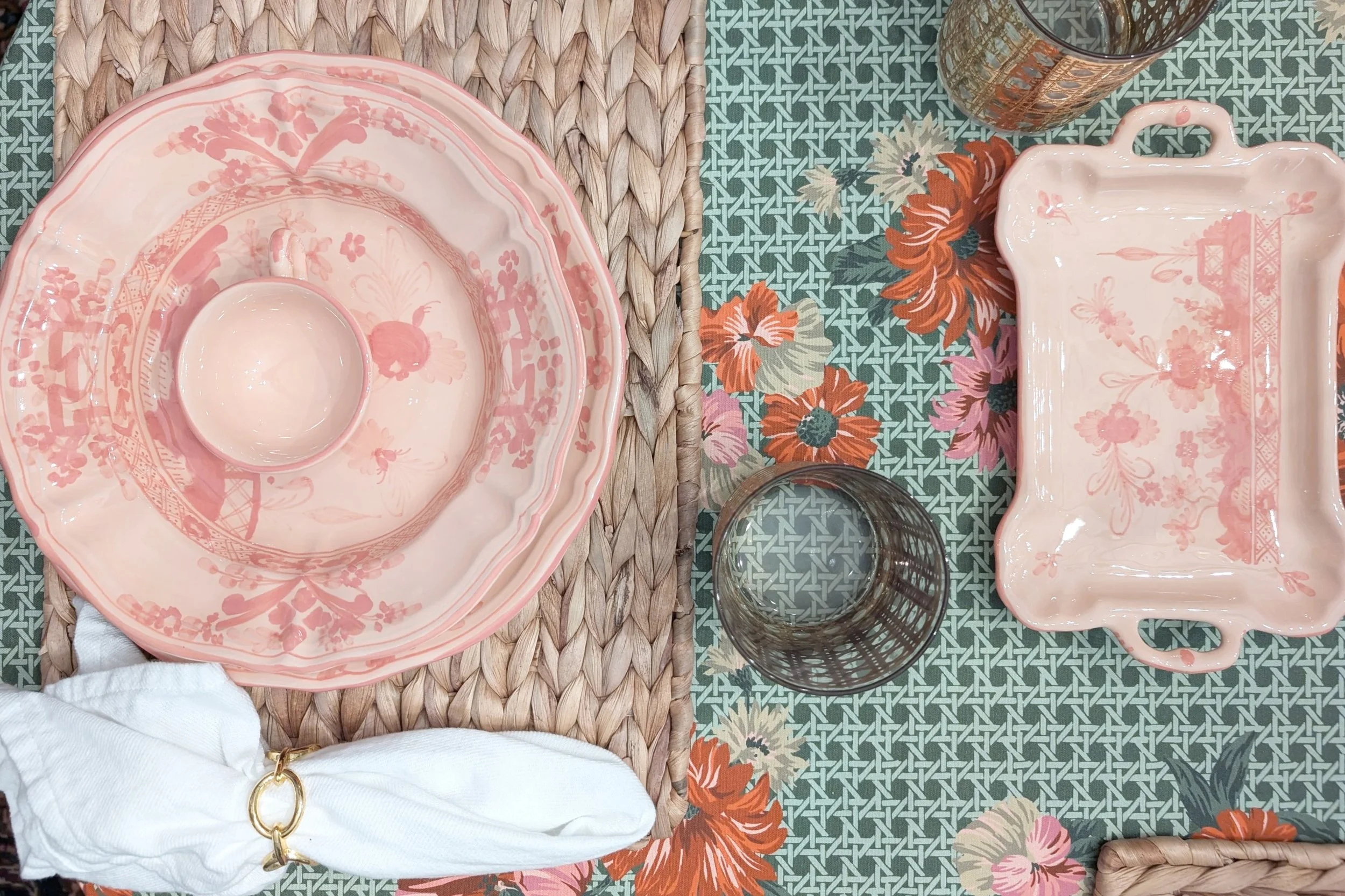 A table setting with pink and floral dishware, including a large pink plate with a bowl in the center, a pink rectangular platter, two transparent glasses, and a floral tablecloth with a woven mat underneath.