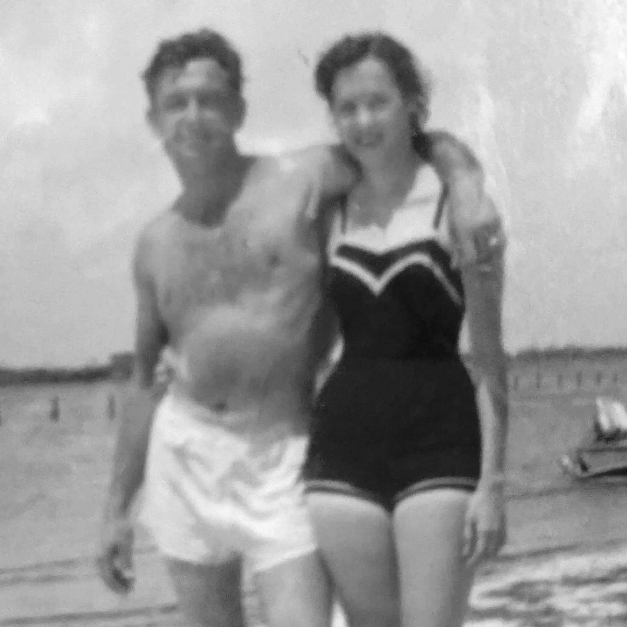 A black and white photo of a young man and woman standing close together outdoors, smiling, with the ocean in the background.