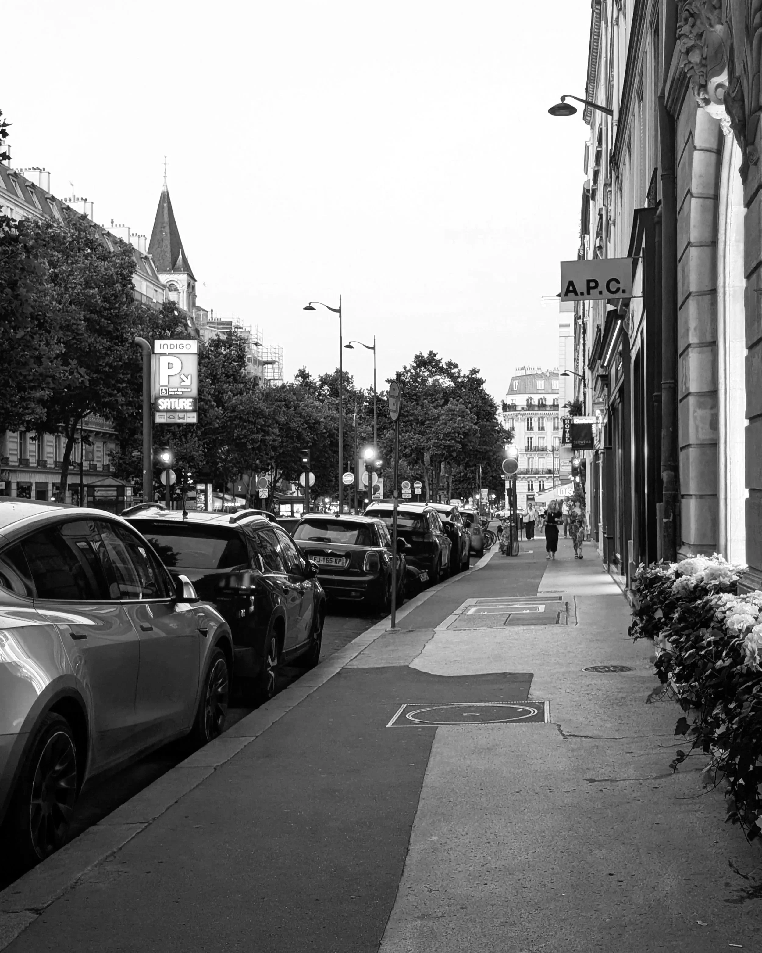 A black-and-white street scene with parked cars along the sidewalk, trees lining the street, and pedestrians walking in the distance. Buildings with signs, including one reading A.P.C., and a church steeple visible in the background.