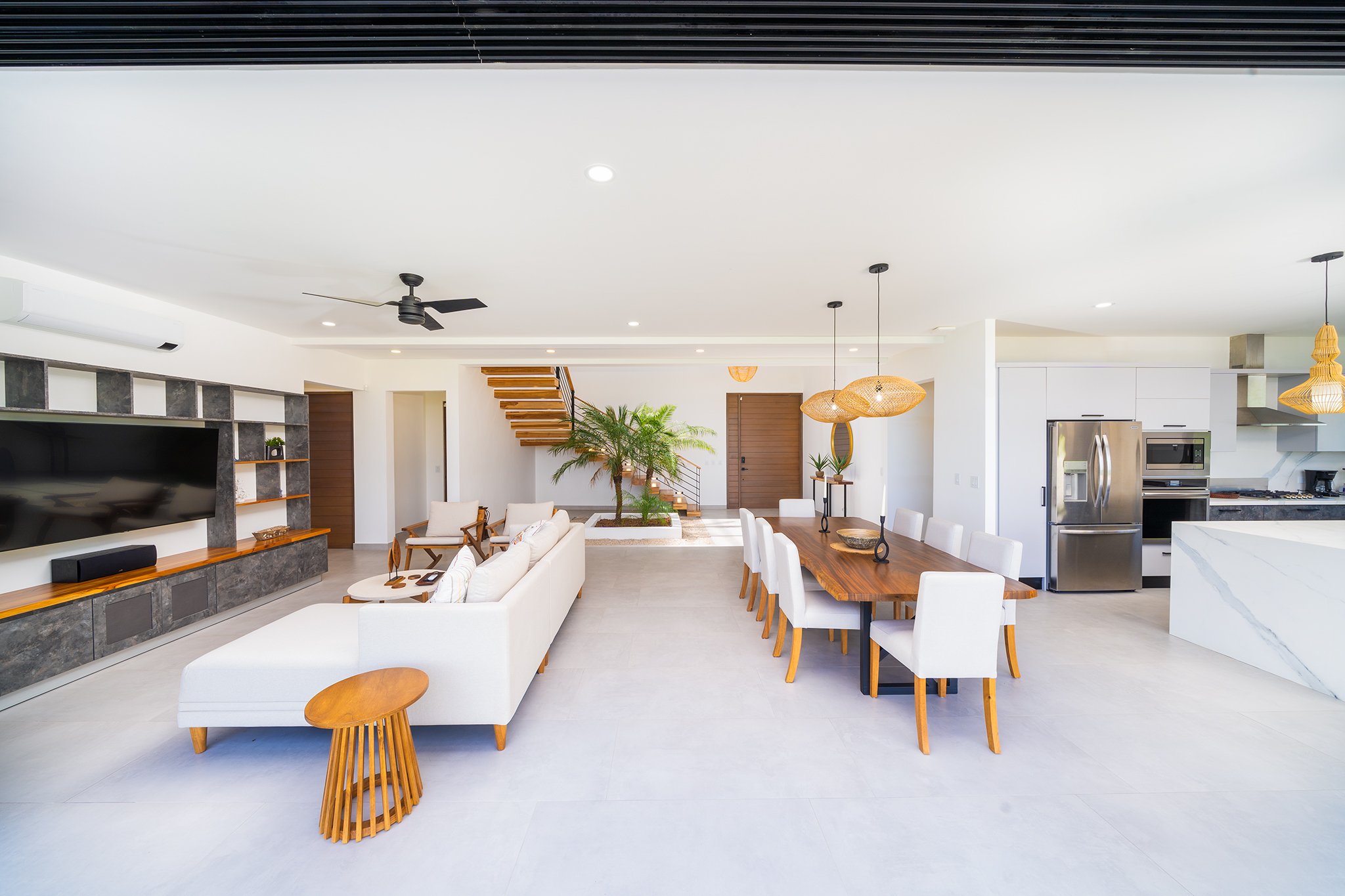 Modern open-concept living room and kitchen with white walls, wooden furniture, a staircase, potted plants, and pendant lights.
