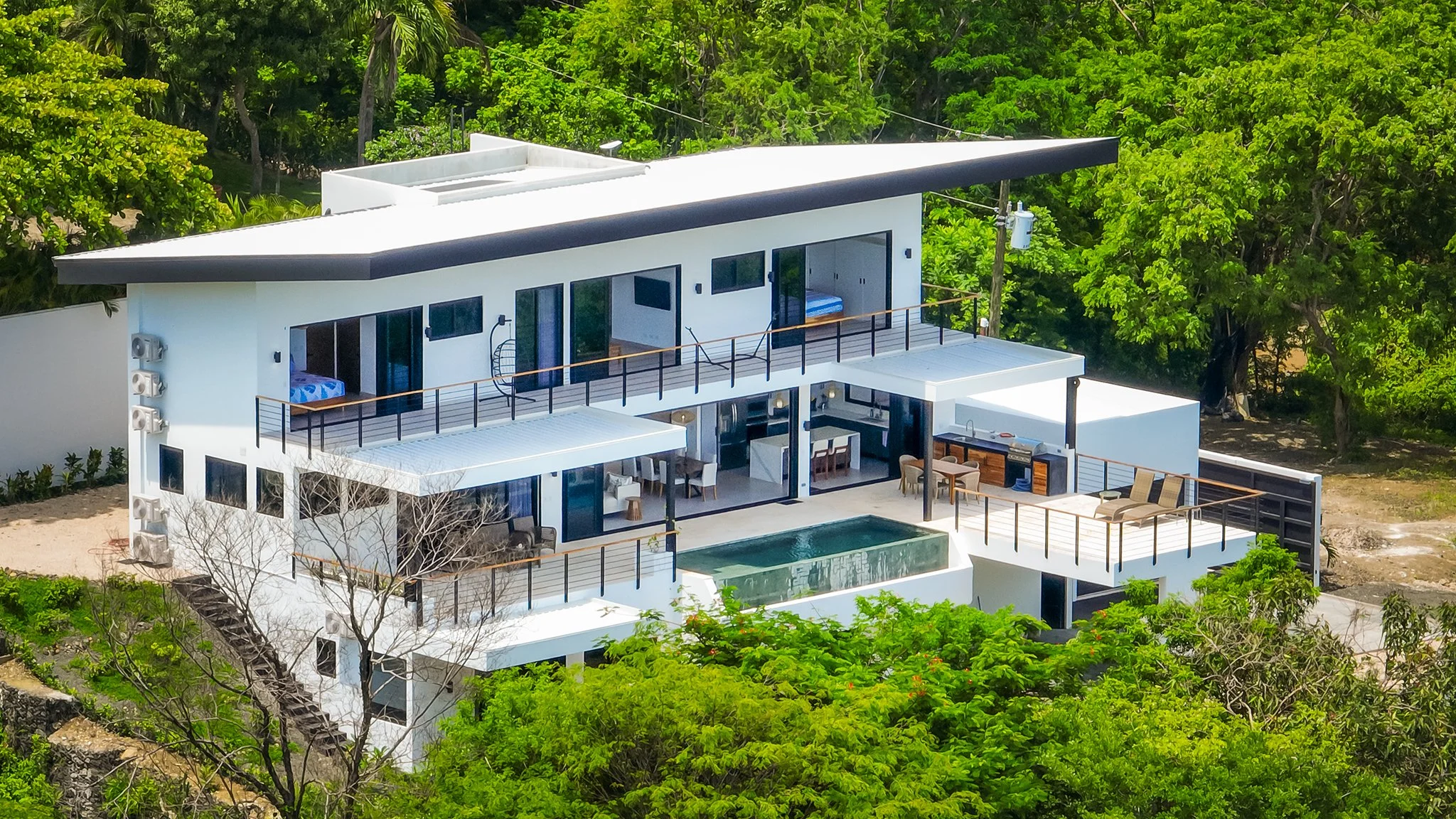 Modern white multi-level house with flat roofs, large glass windows, and outdoor terraces surrounded by lush green trees.
