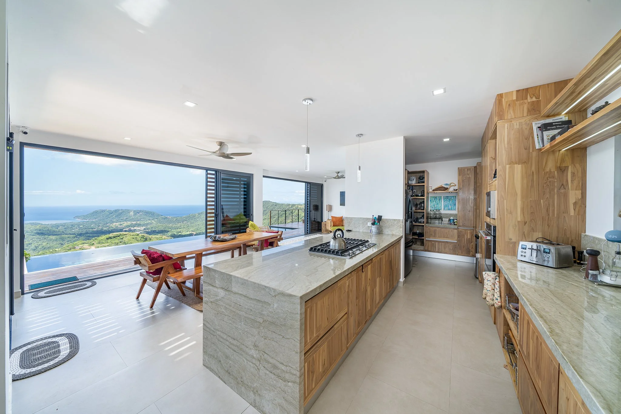 Modern open-concept kitchen with a view of lush green hills and ocean through large glass sliding doors, featuring wooden cabinets, a kitchen island, and minimalist decor.