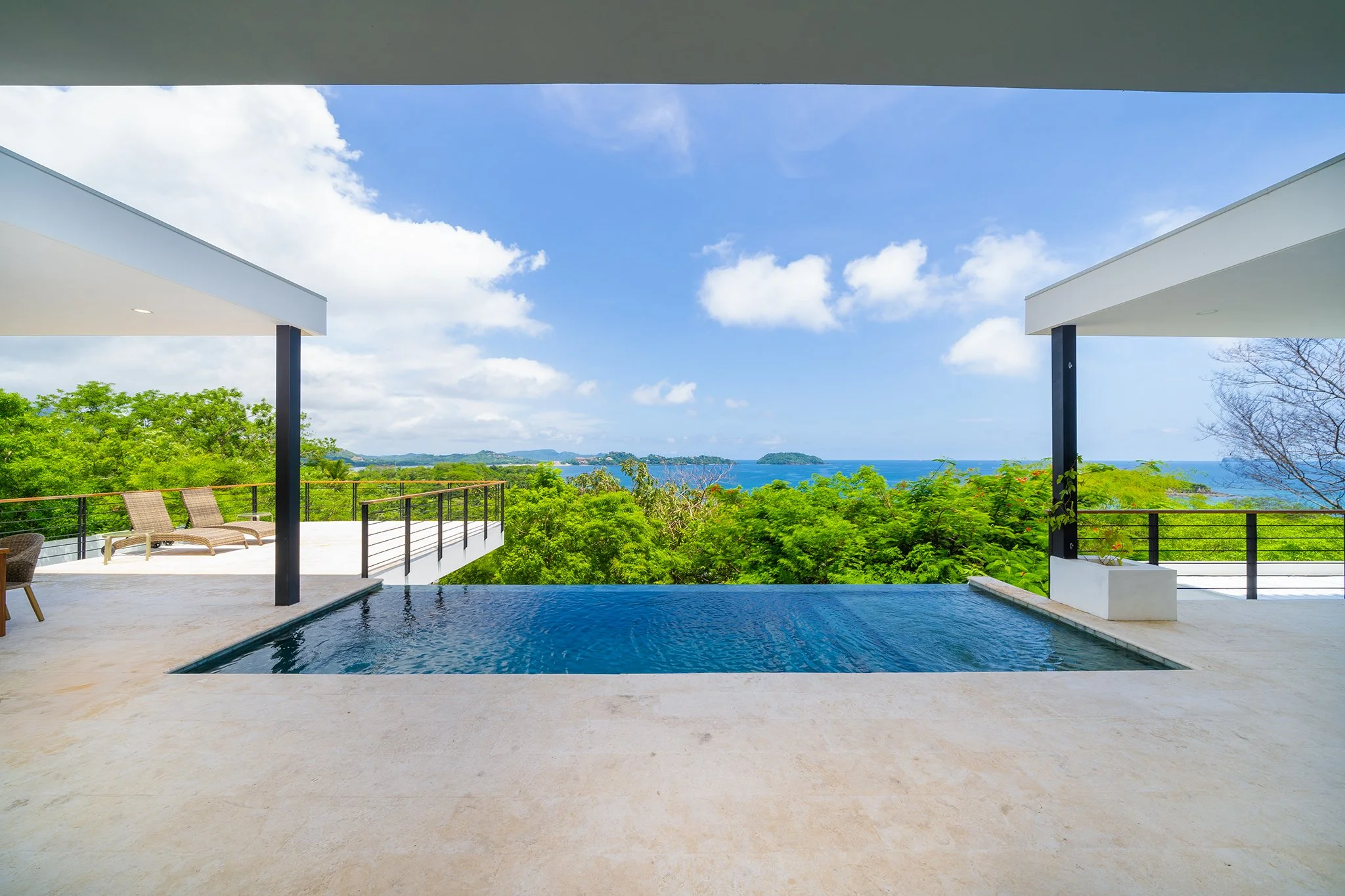 View of a swimming pool with lounge chairs and a railing, overlooking lush green trees and blue ocean in the distance under a partly cloudy sky, framed by the edges of a white modern building.