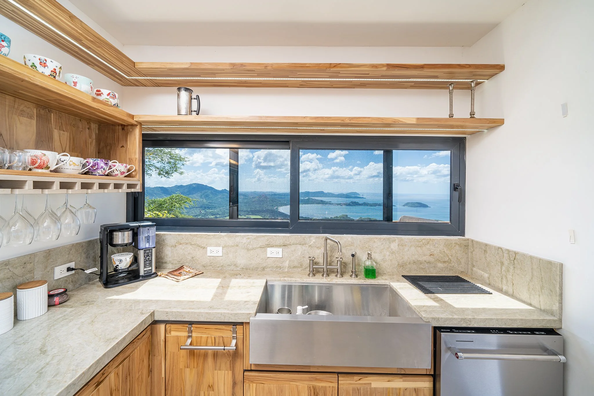 A modern kitchen with a large window showing mountain and ocean views, wooden open shelves, a granite countertop, a small coffee machine, and colorful cups.