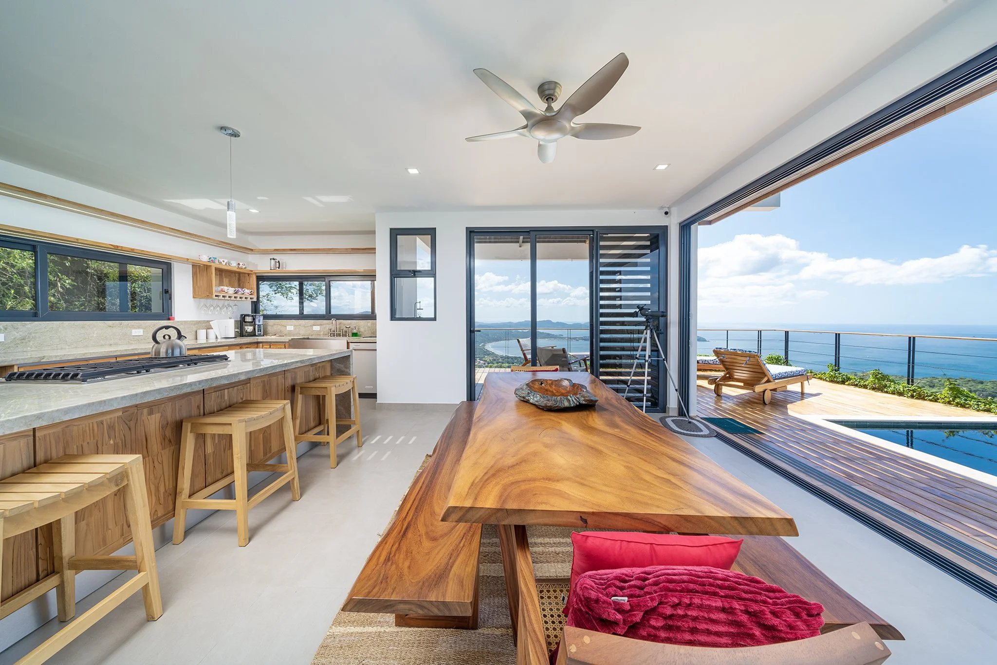 Open-concept kitchen and dining area with large sliding glass doors leading to a balcony with ocean view, modern ceiling fan, island with bar stools, and indoor-outdoor living space.