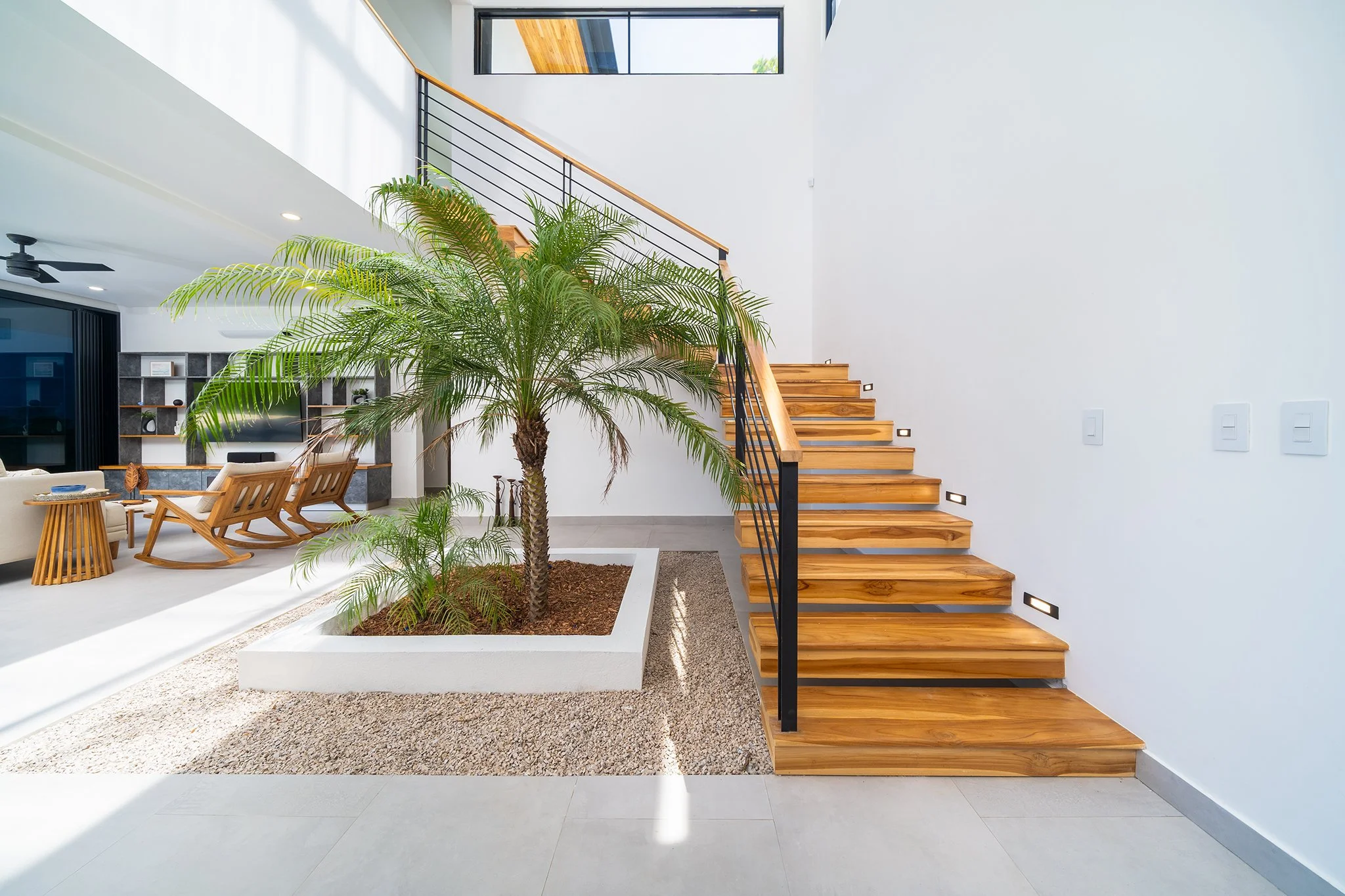 Modern interior with a large indoor palm tree in a white planter, wooden staircase with black metal railing, and a seating area with wooden chairs in a bright, open space with white walls and large windows.