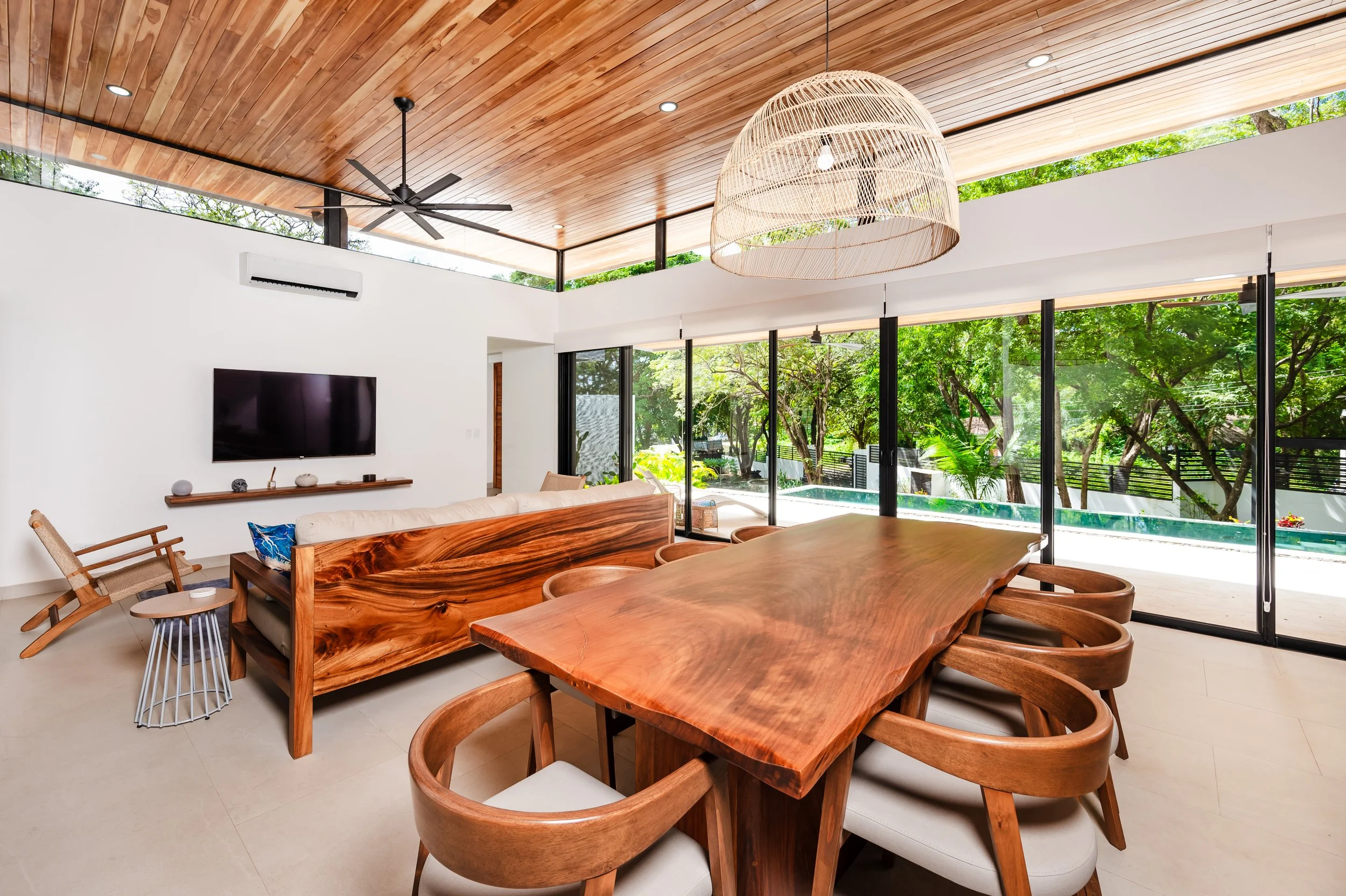 Modern living room with wooden dining table, matching chairs, and a long wooden sofa with navy and white pillows. Large windows reveal a lush green outdoor scenery with trees and a pool. The ceiling features wood paneling, a black ceiling fan, and a 