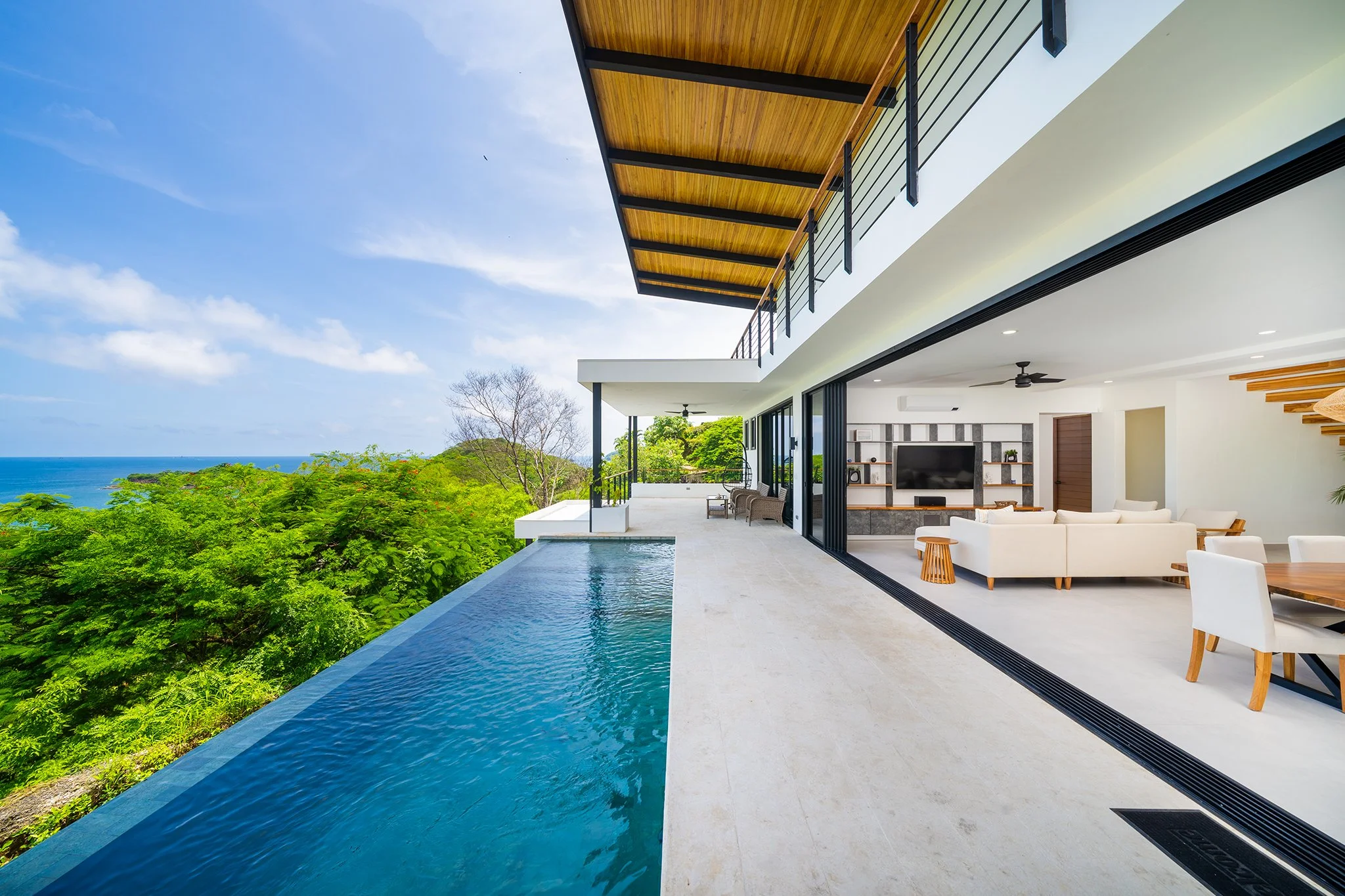 Modern house with open living room, large glass sliding doors, white furniture, and a pool overlooking lush green trees and ocean view under a clear blue sky.